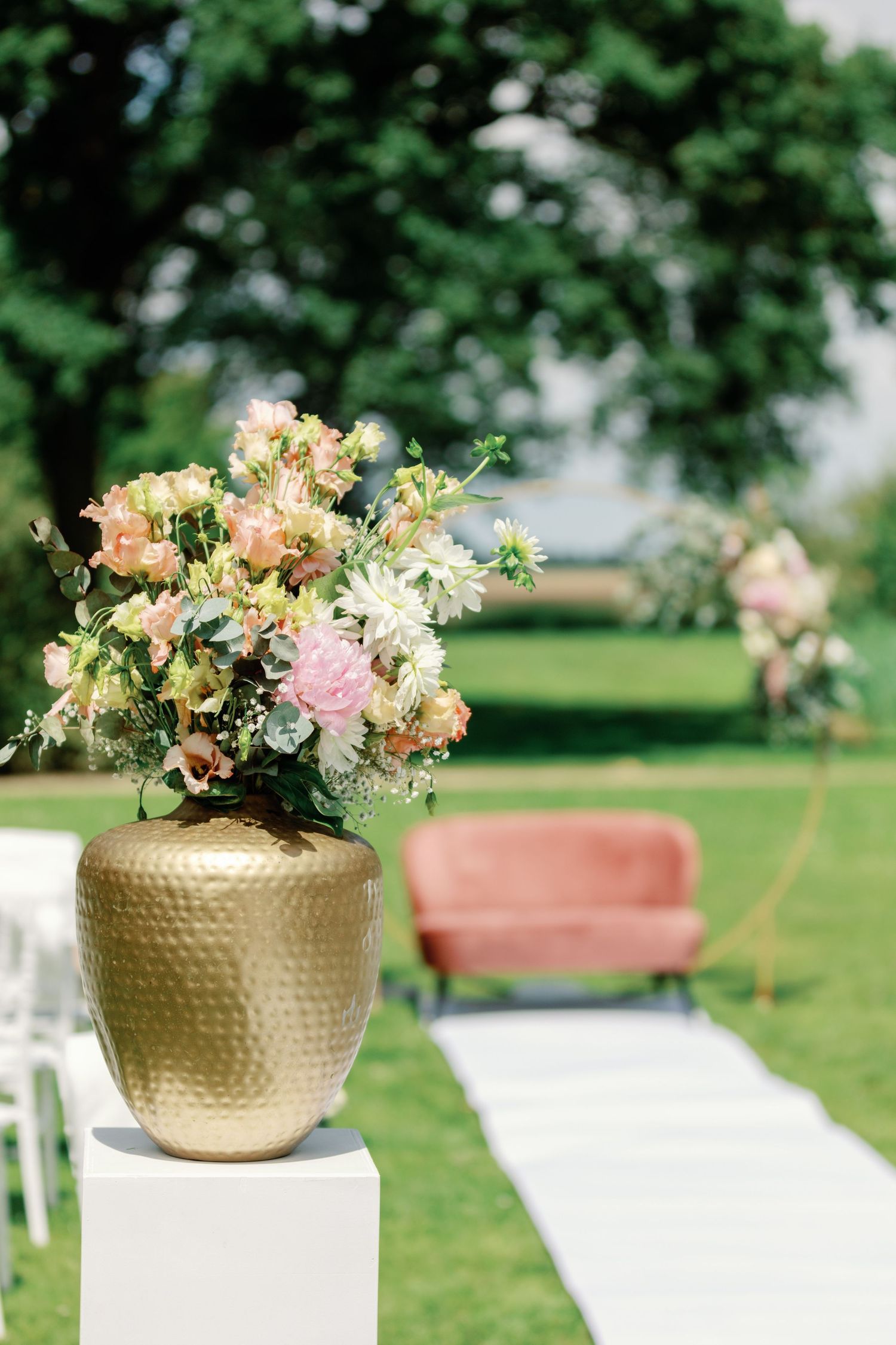 Elegant gold metallic vase filled with pink and cream flowers sits on white pedestal at outdoor garden event.