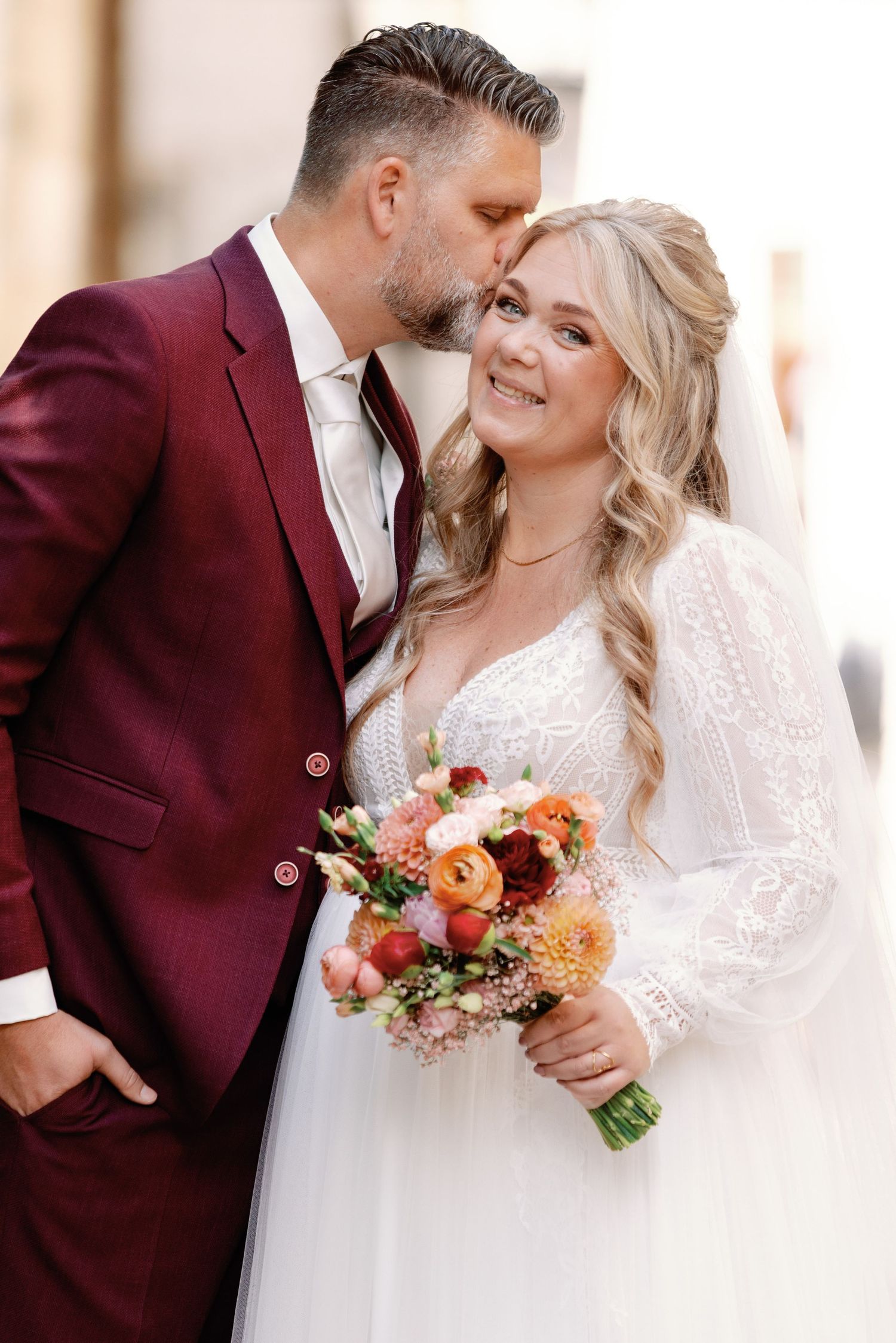 Romantic wedding couple embraces in burgundy suit and white lace gown with colorful autumn bouquet.