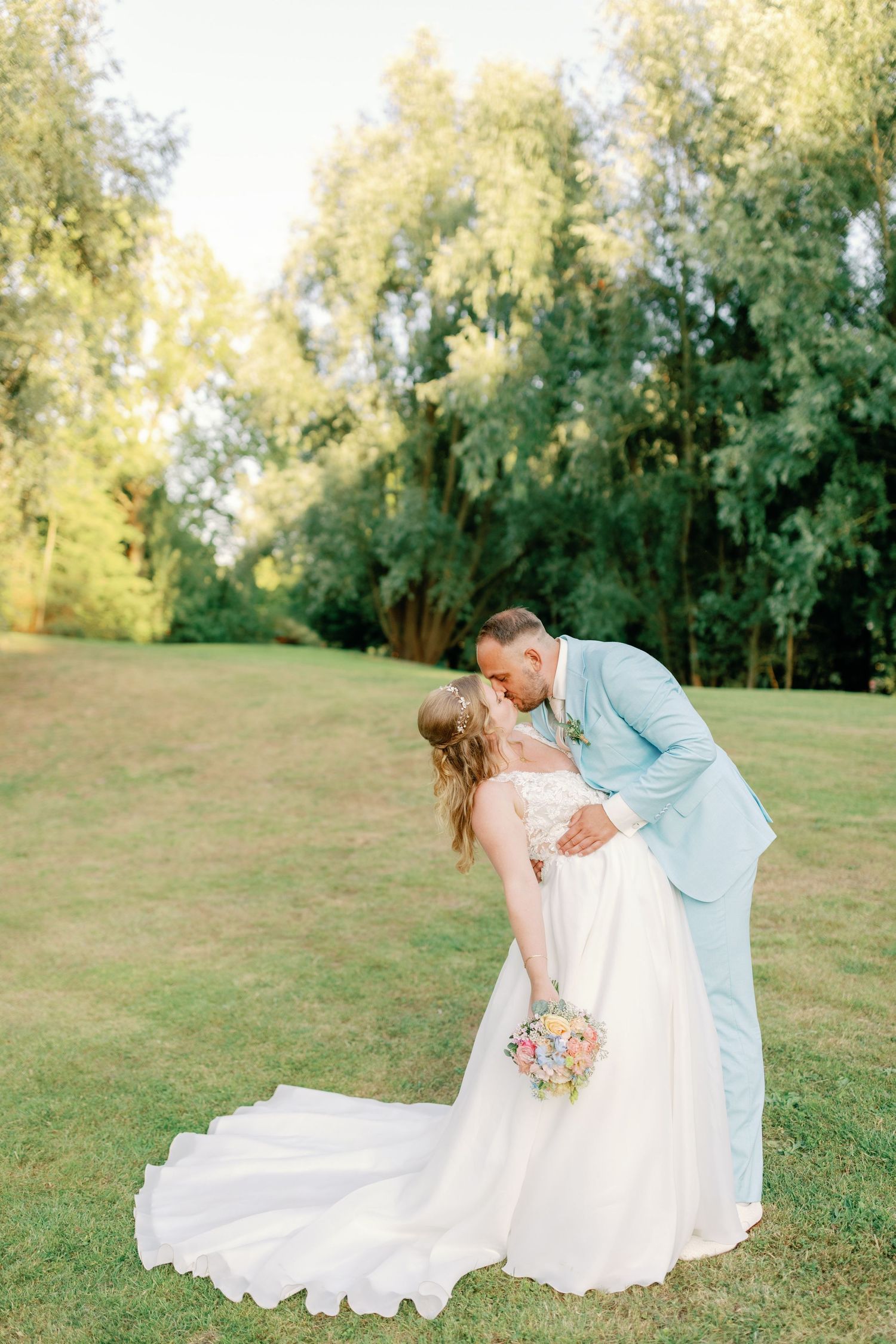 A romantic wedding couple shares a kiss in a lush garden setting with flowing white dress and light blue suit.