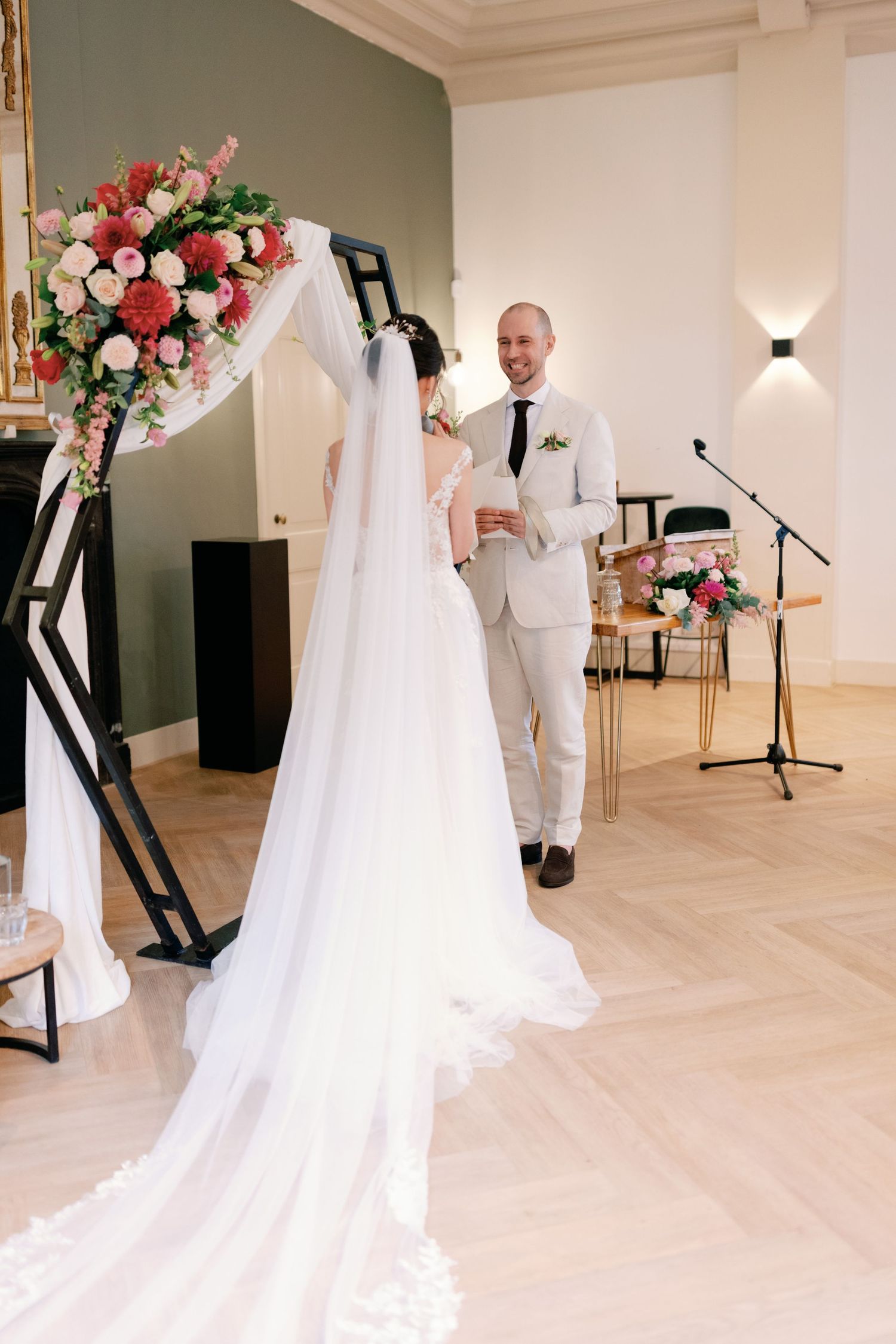 An elegant wedding ceremony setup with a floral arch and long flowing white veil against sage green walls.