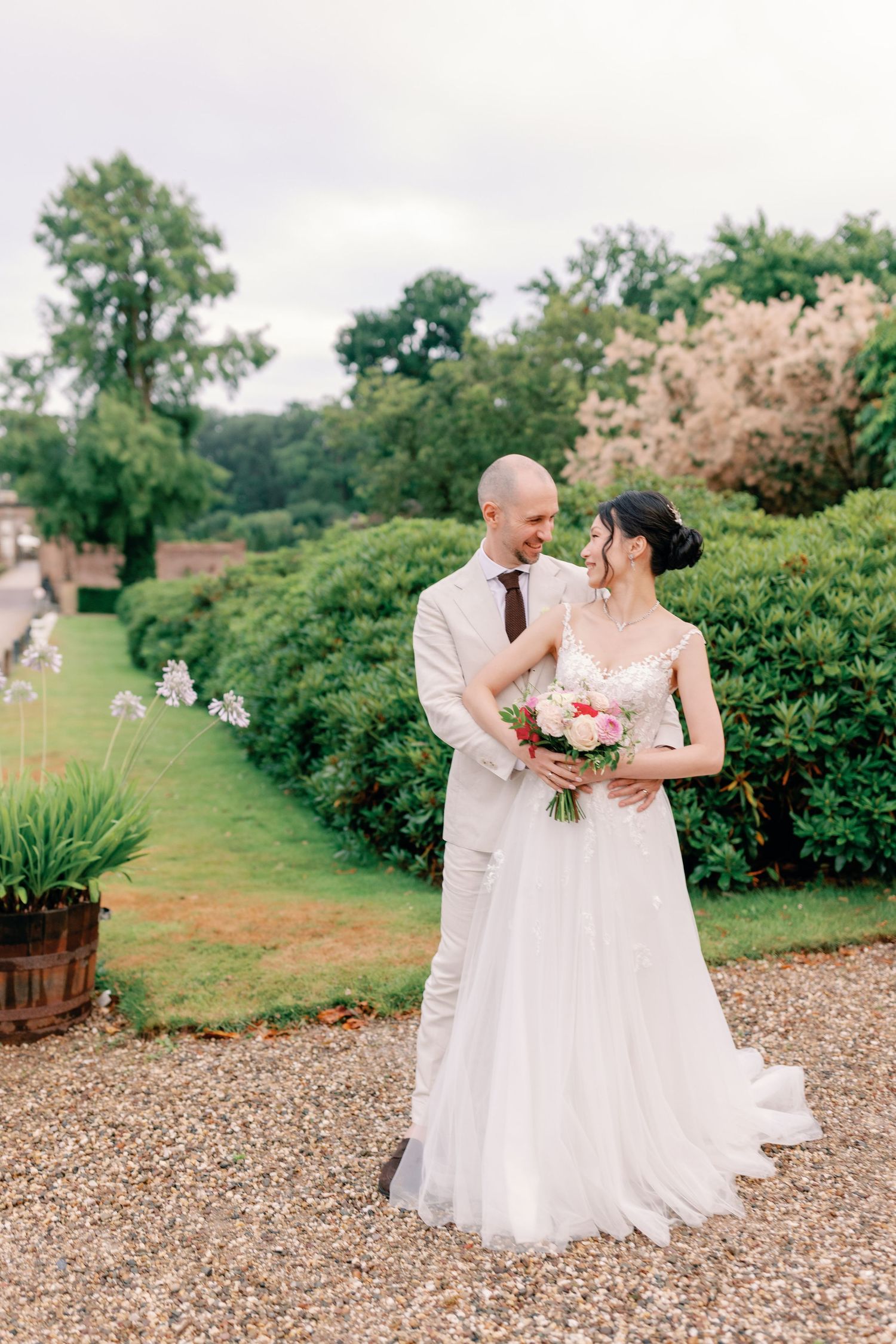 A romantic wedding couple shares a tender moment on a gravel path surrounded by lush summer gardens.