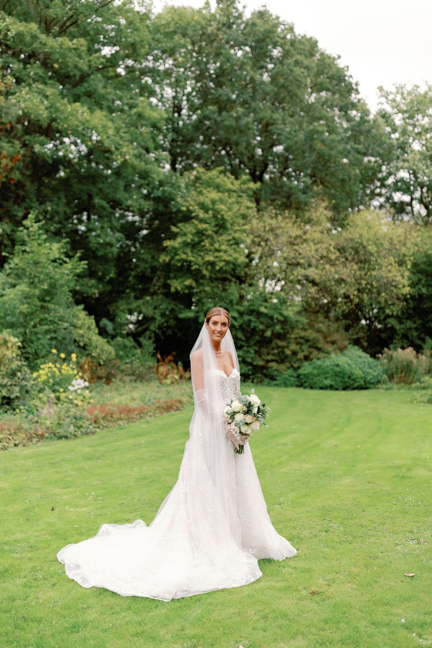 A bride in a flowing white lace gown with long train poses on a manicured lawn surrounded by lush greenery.