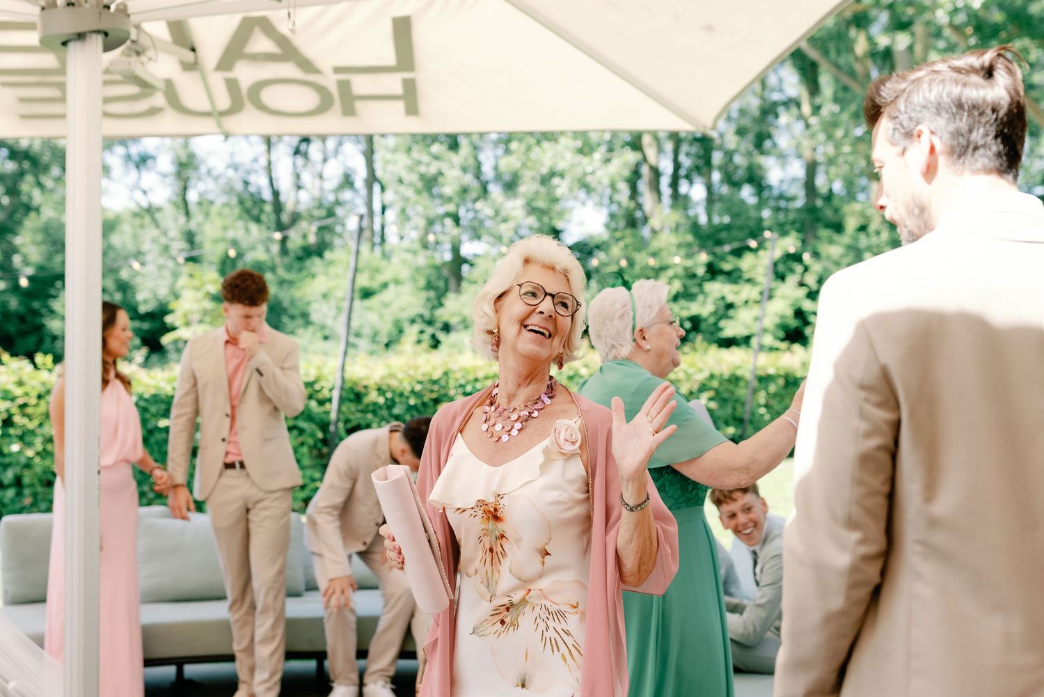 A wedding party poses together outdoors wearing pastel green dresses and light beige suits in a garden setting.