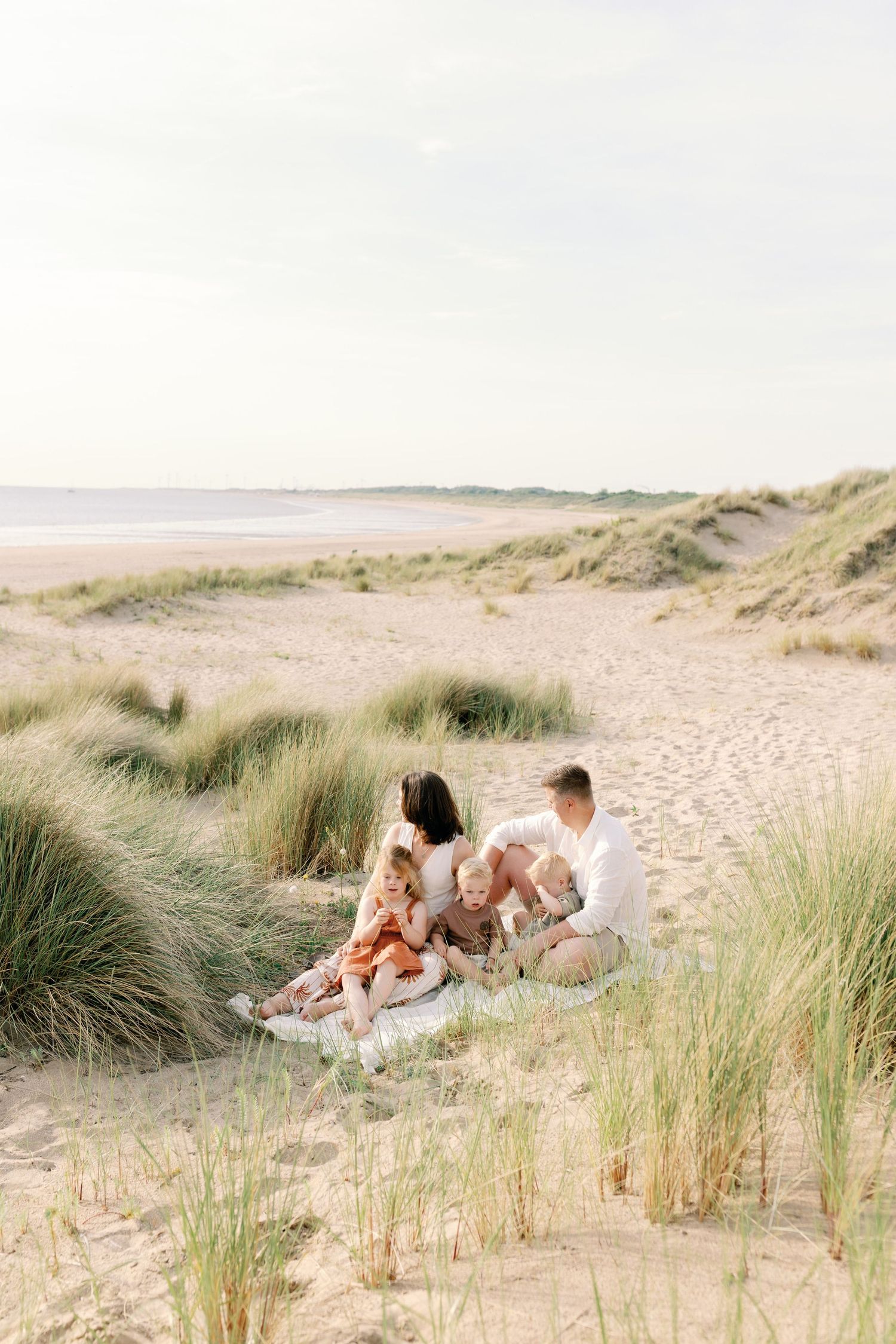 Family of four sitting on a blanket among sand dunes at the beach, enjoying a sunny day together.