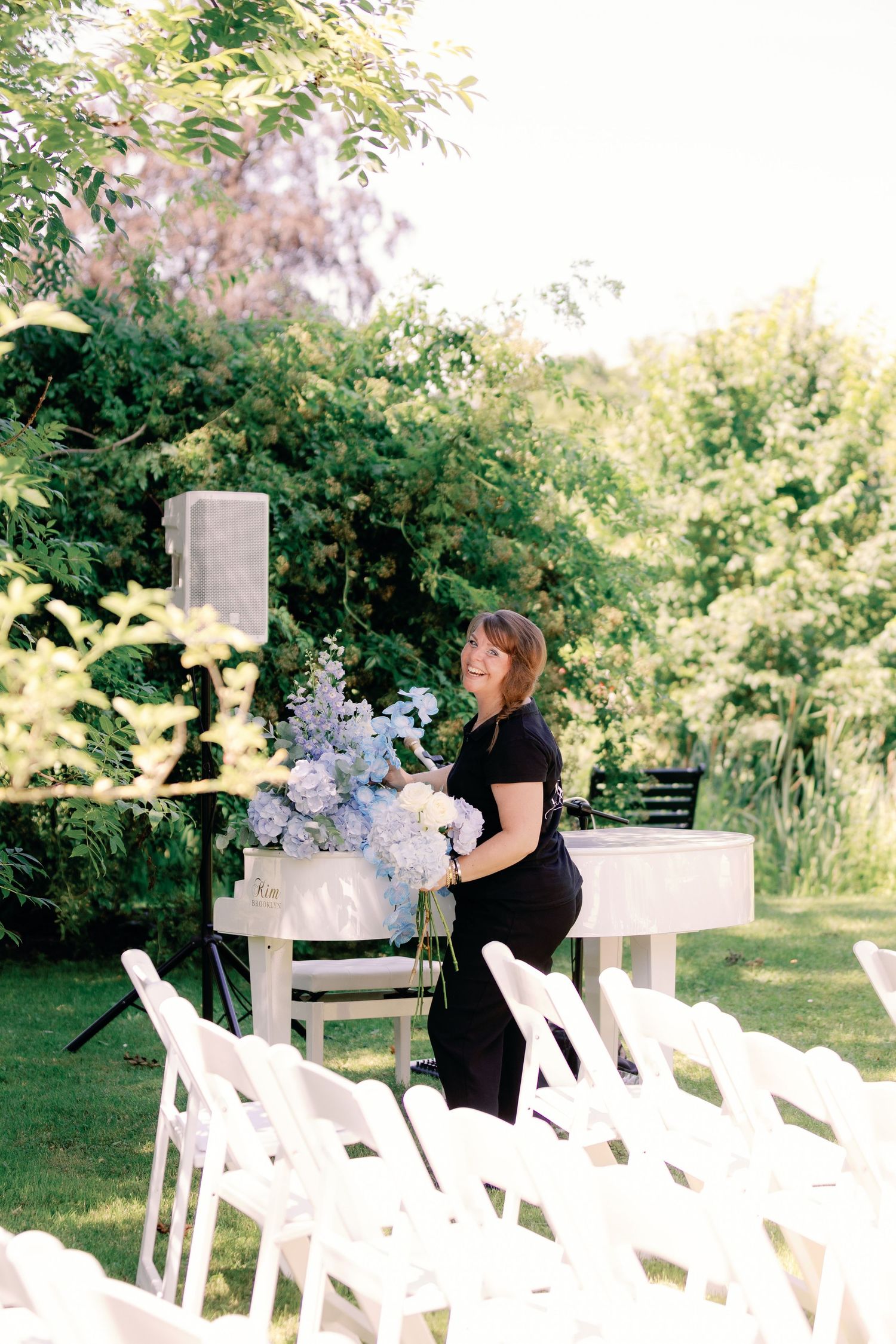Woman in black dress arranging blue hydrangeas at an outdoor wedding ceremony with white chairs.