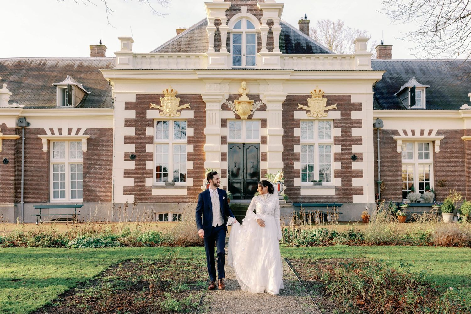 A couple in wedding attire stands in front of an ornate historic brick mansion with decorative architectural details.