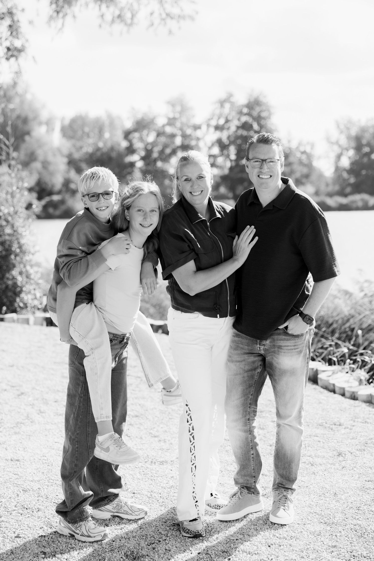 Black and white family portrait of two adults and two children smiling and embracing outdoors on a lawn beside a lake with trees behind.