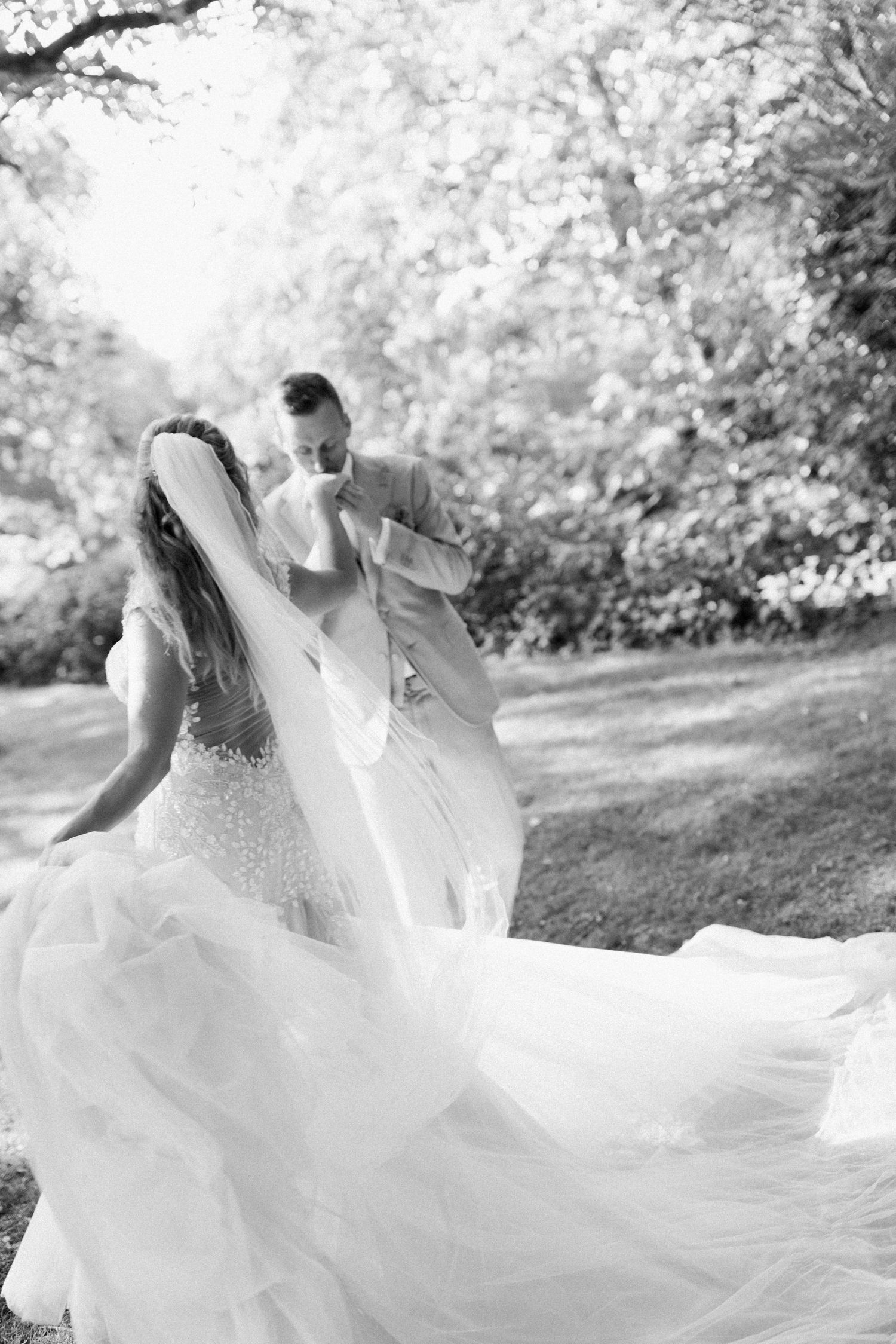 Black and white photo of a bride and groom dancing outdoors, her flowing gown and veil billowing in the breeze.