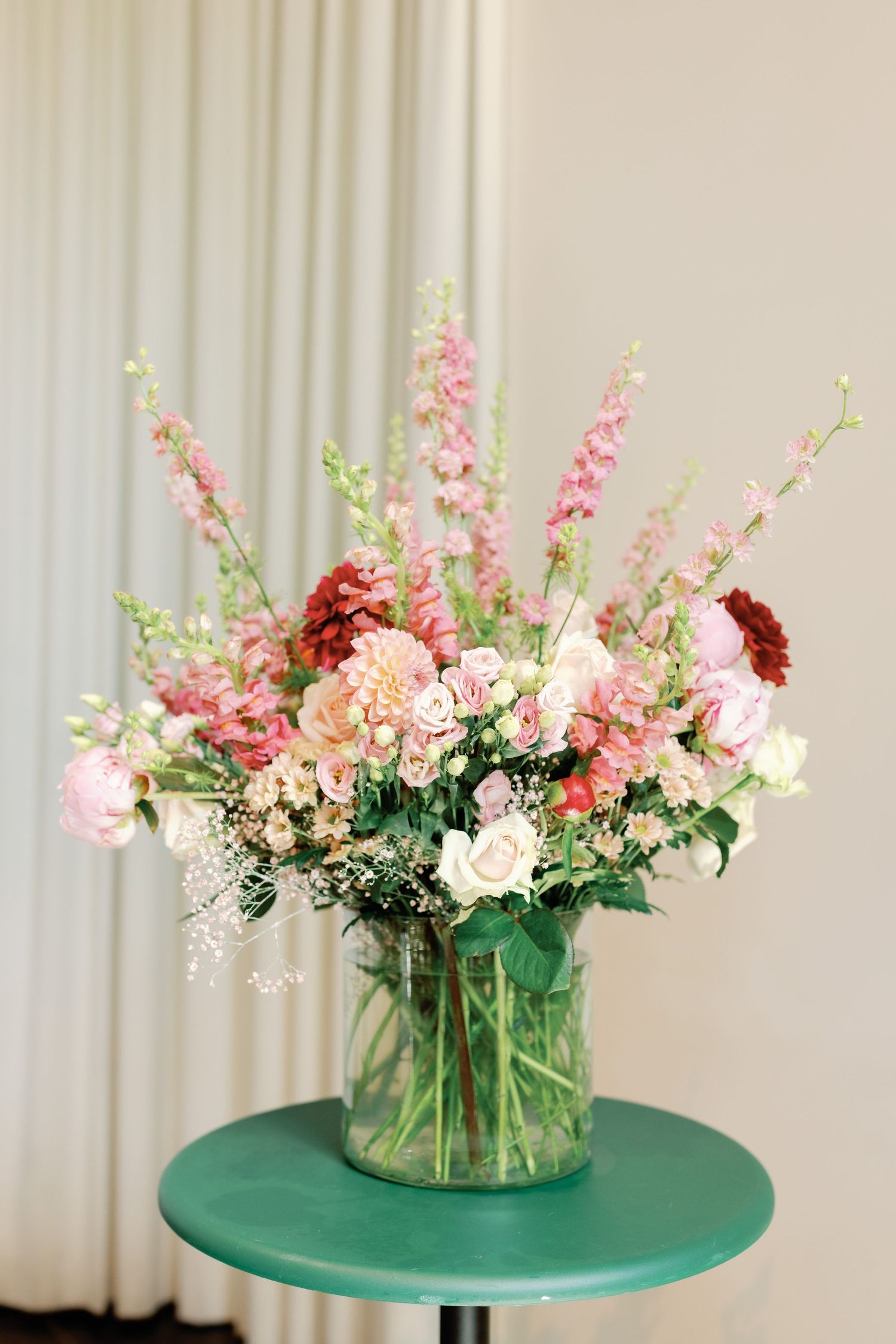 Pink and white floral arrangement in clear glass vase displayed on turquoise table with white curtain backdrop.