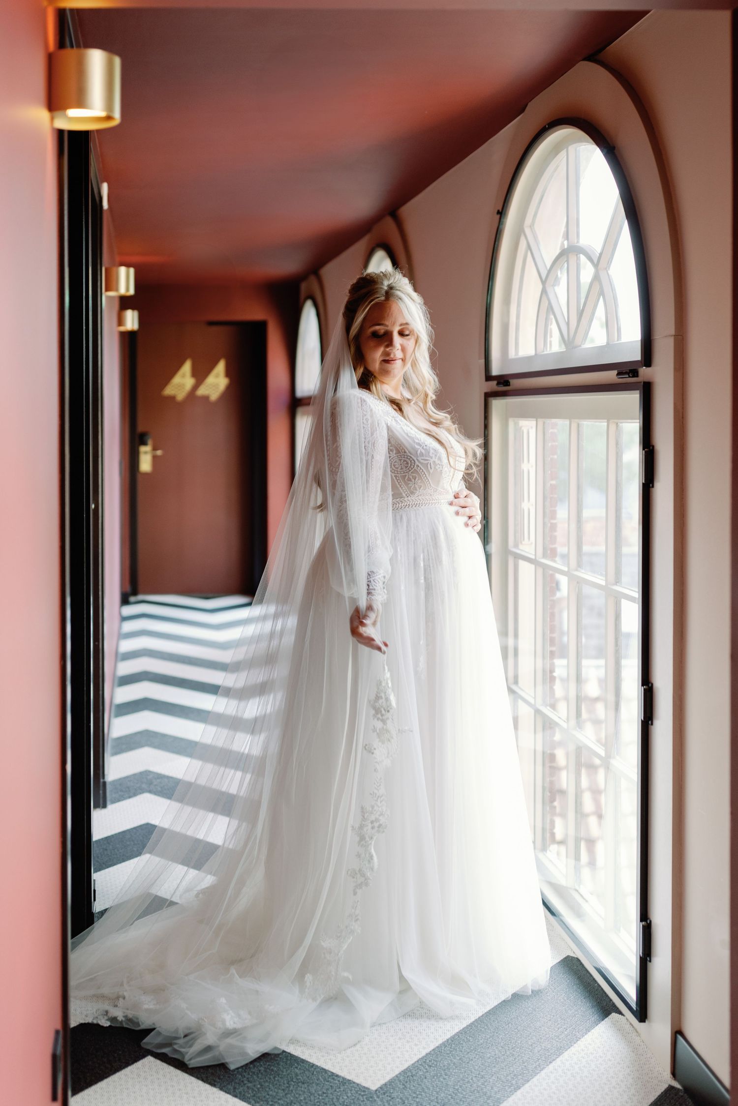 A bride in an elegant white wedding dress and veil poses by an arched window with natural light streaming in.