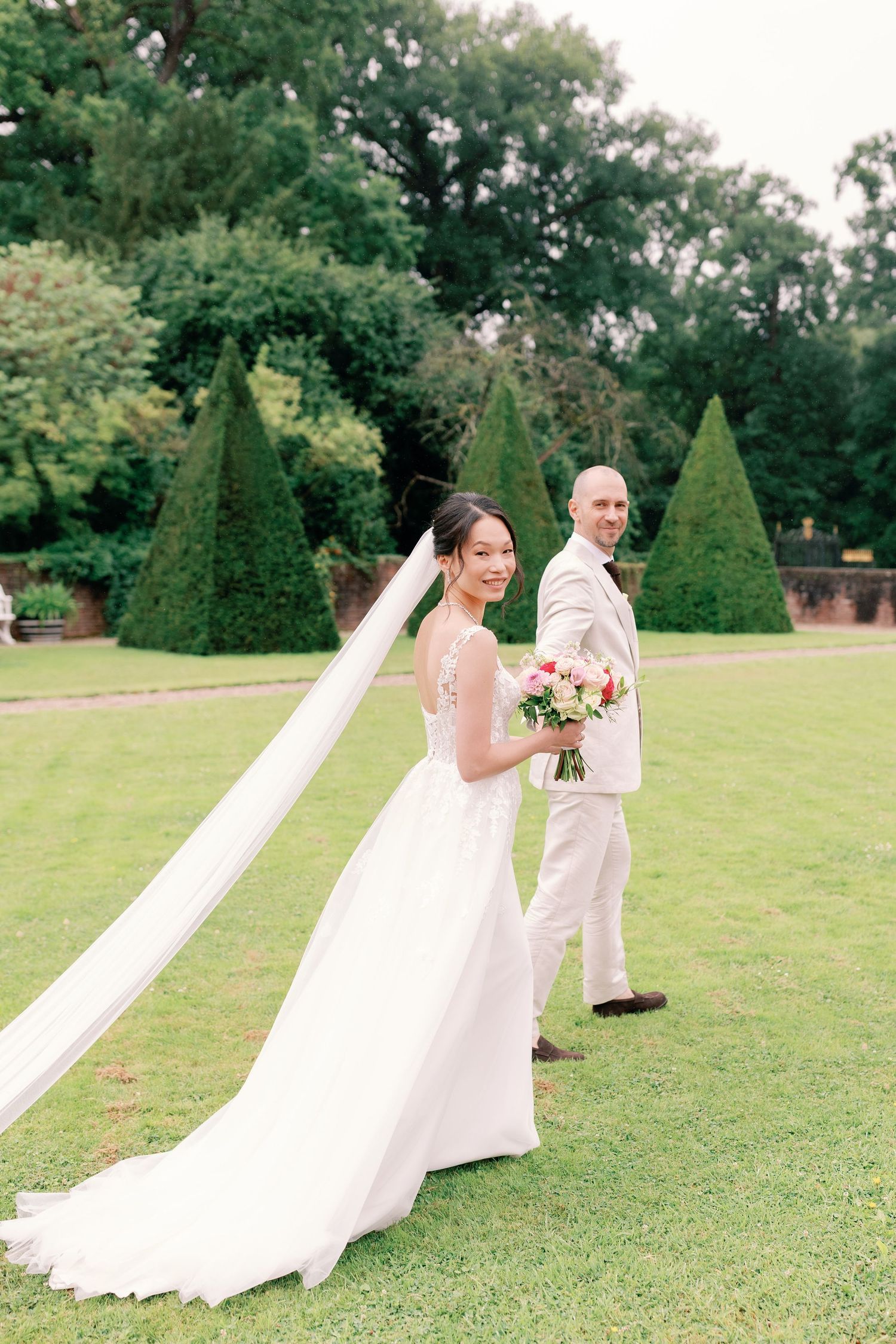 Bride and groom walk hand in hand across a manicured lawn with ornamental topiary trees in the background.