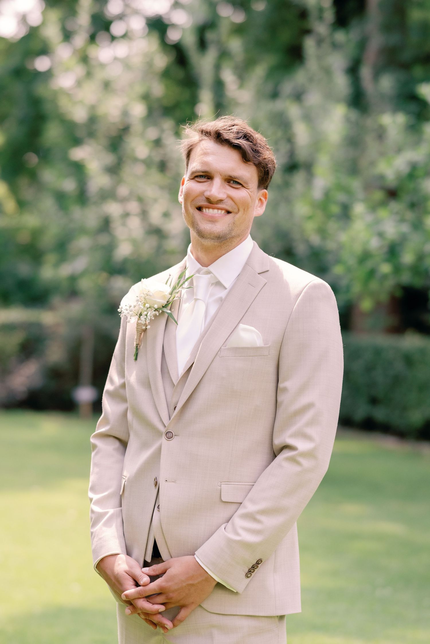 A groom in a light beige suit and pink tie smiles while standing outdoors in a garden setting.