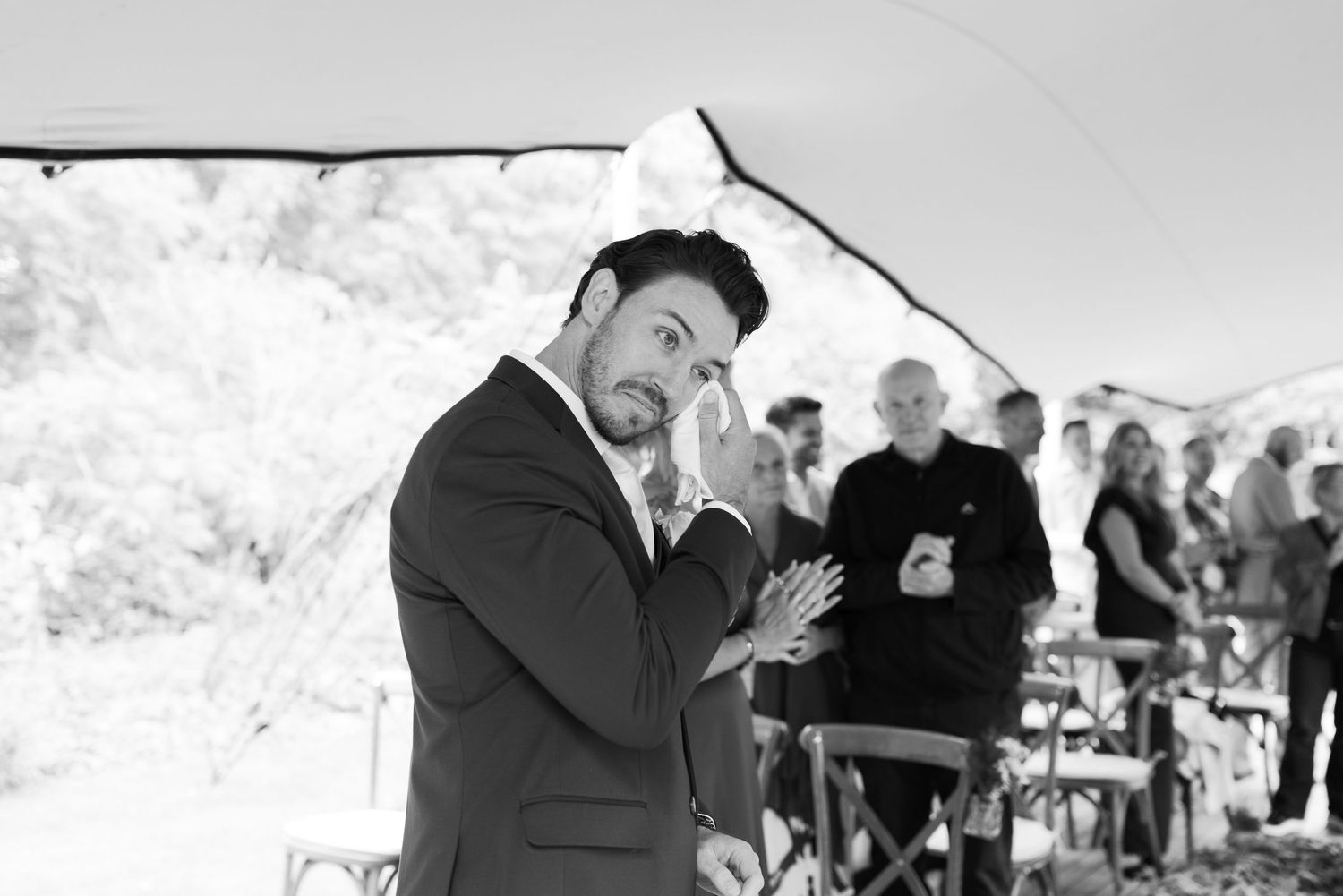 Black and white photo of an emotional groom reacting during an outdoor wedding ceremony under a tent.
