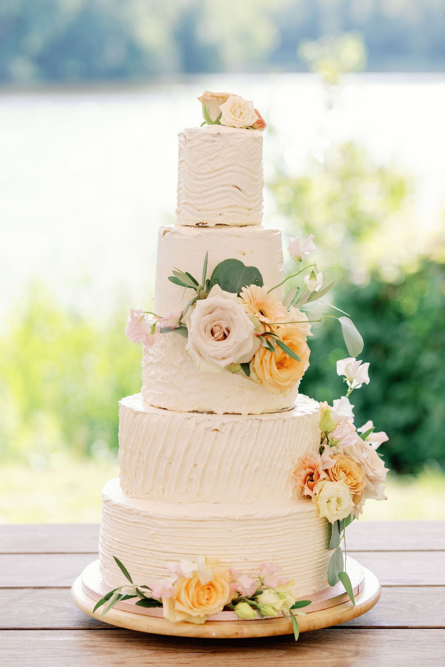 Three-tiered white wedding cake with peach and cream roses and greenery decorations on a wooden table.