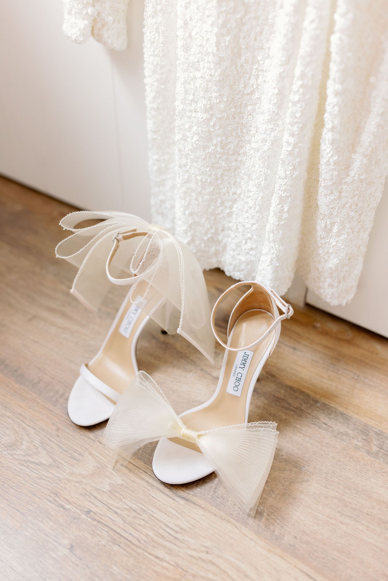 White bridal heels with ankle straps displayed against wooden floor and lace curtain.