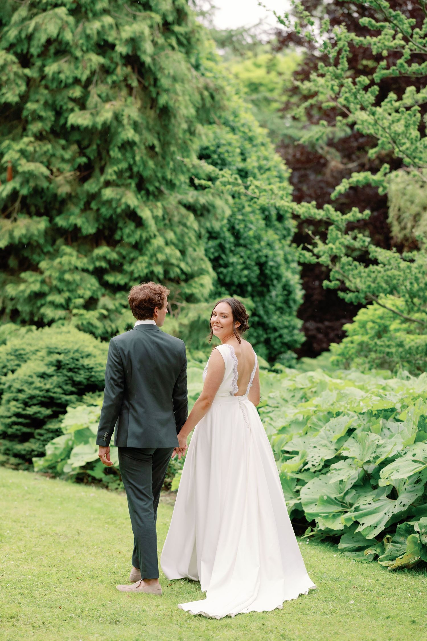 Couple in wedding attire walking through a lush green garden setting.