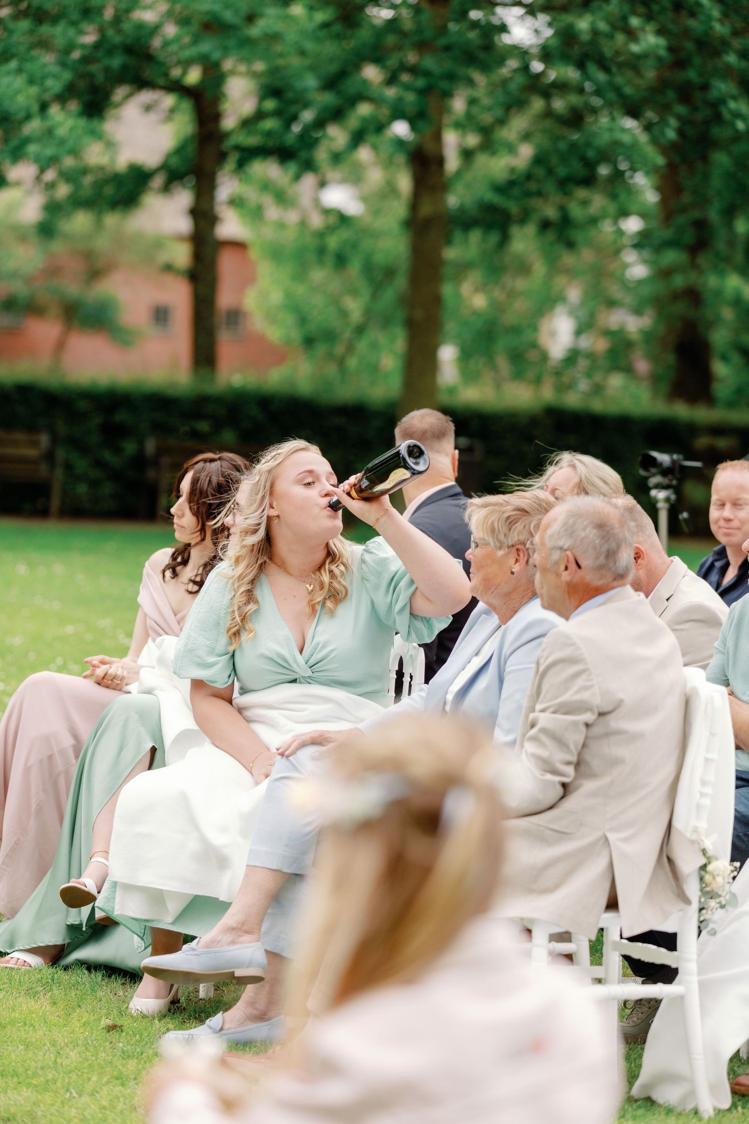 Wedding guests sitting in white chairs outdoors laugh together during a joyful moment at a garden ceremony.