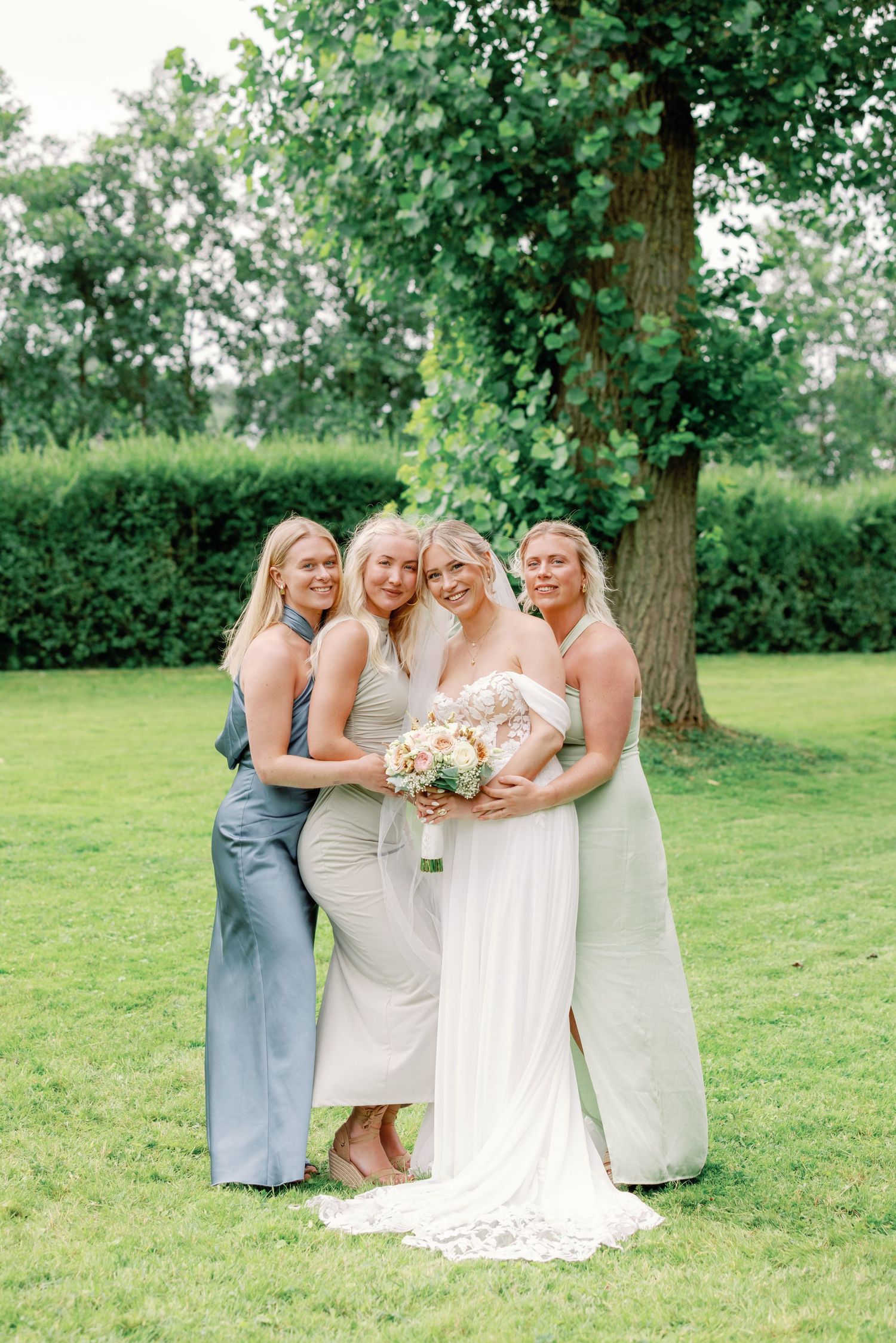 A group of bridesmaids in soft pastel dresses share a candid laughing moment on a green lawn during a wedding.