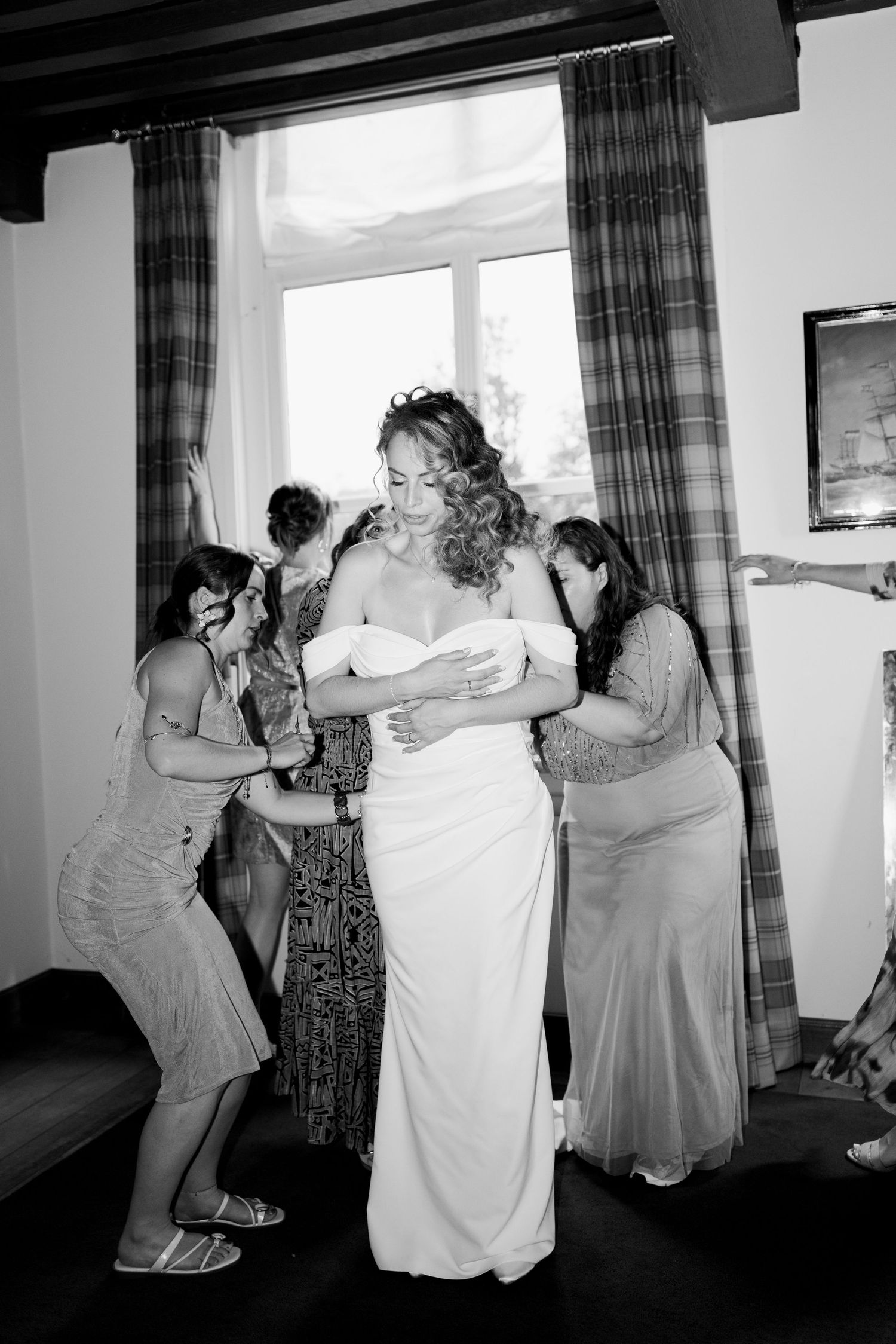 Black and white photo of a bride in a white gown being helped by bridesmaids near a window on her wedding day.