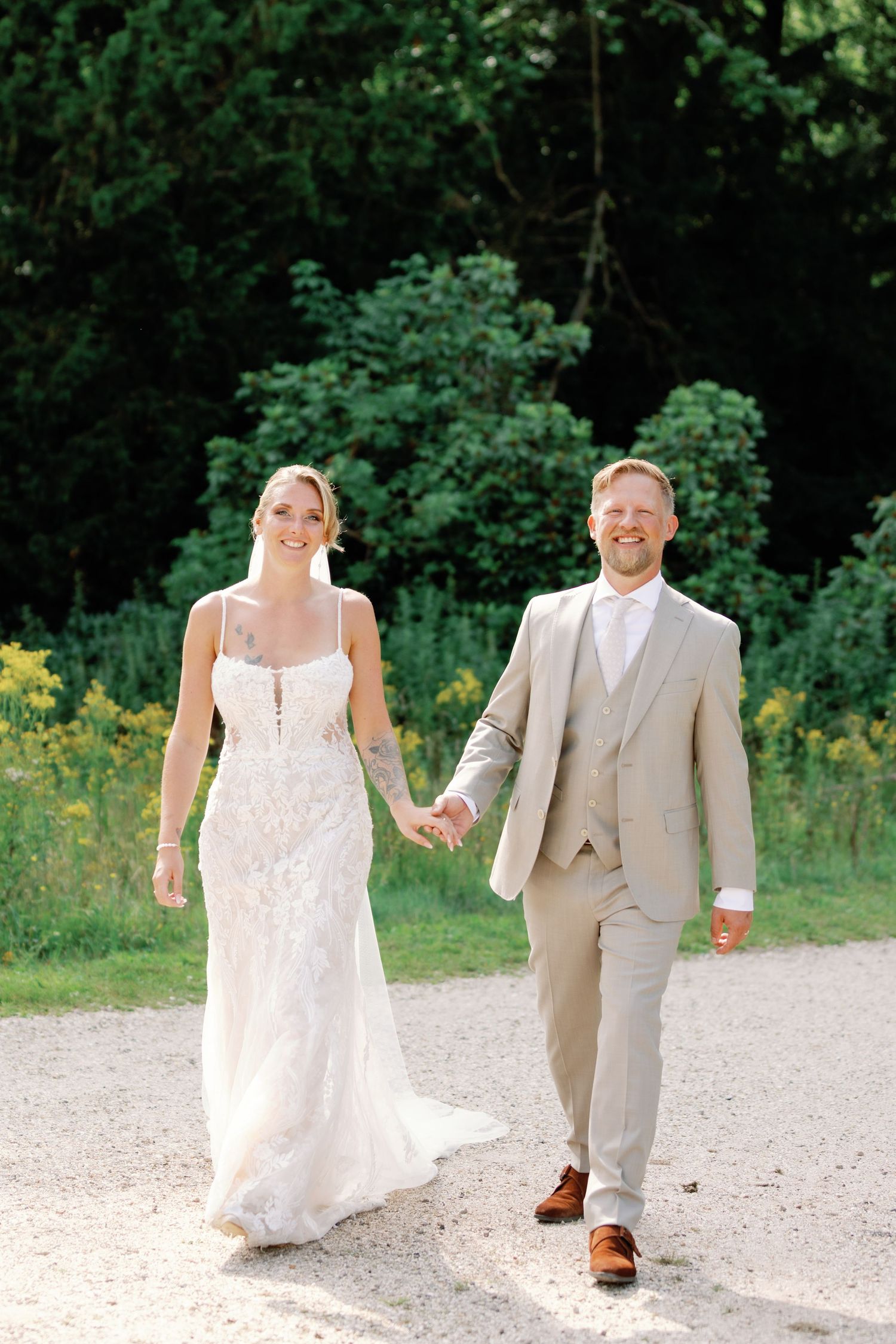 A couple in wedding attire walks and dances playfully on a gravel path surrounded by greenery.