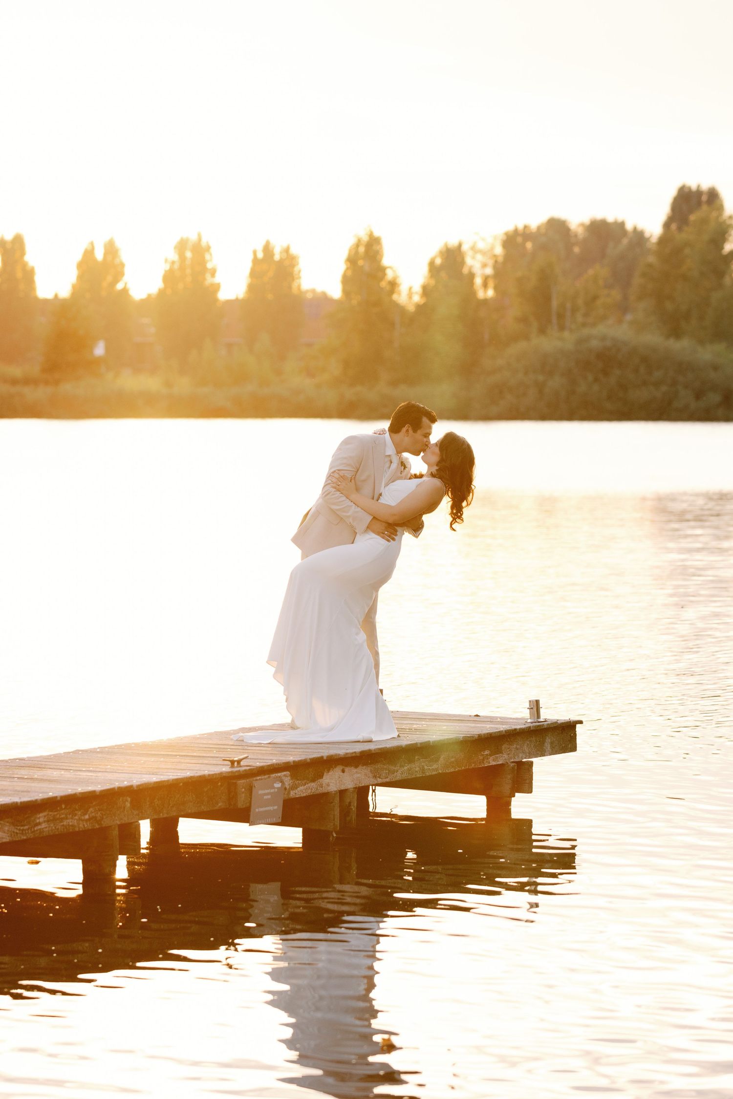 A couple sits romantically on a wooden dock at sunset overlooking a tranquil lake with golden sunlight.