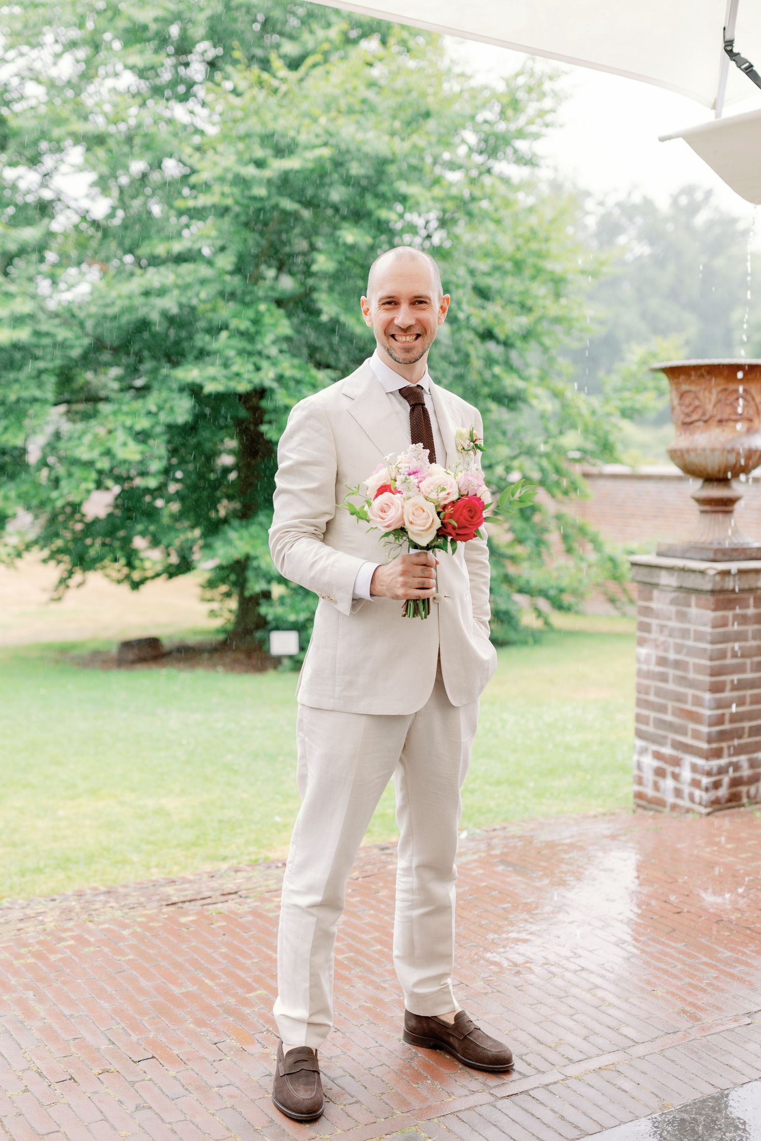 A person in a light beige suit holds a pink and red bouquet while standing on a brick patio surrounded by greenery.