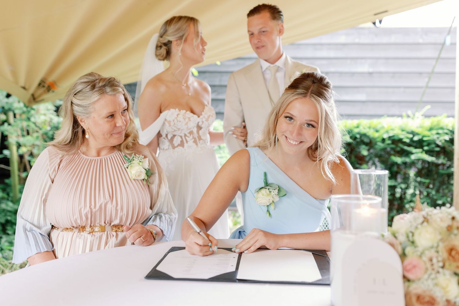 People signing documents at an outdoor wedding ceremony under a cream-colored tent.