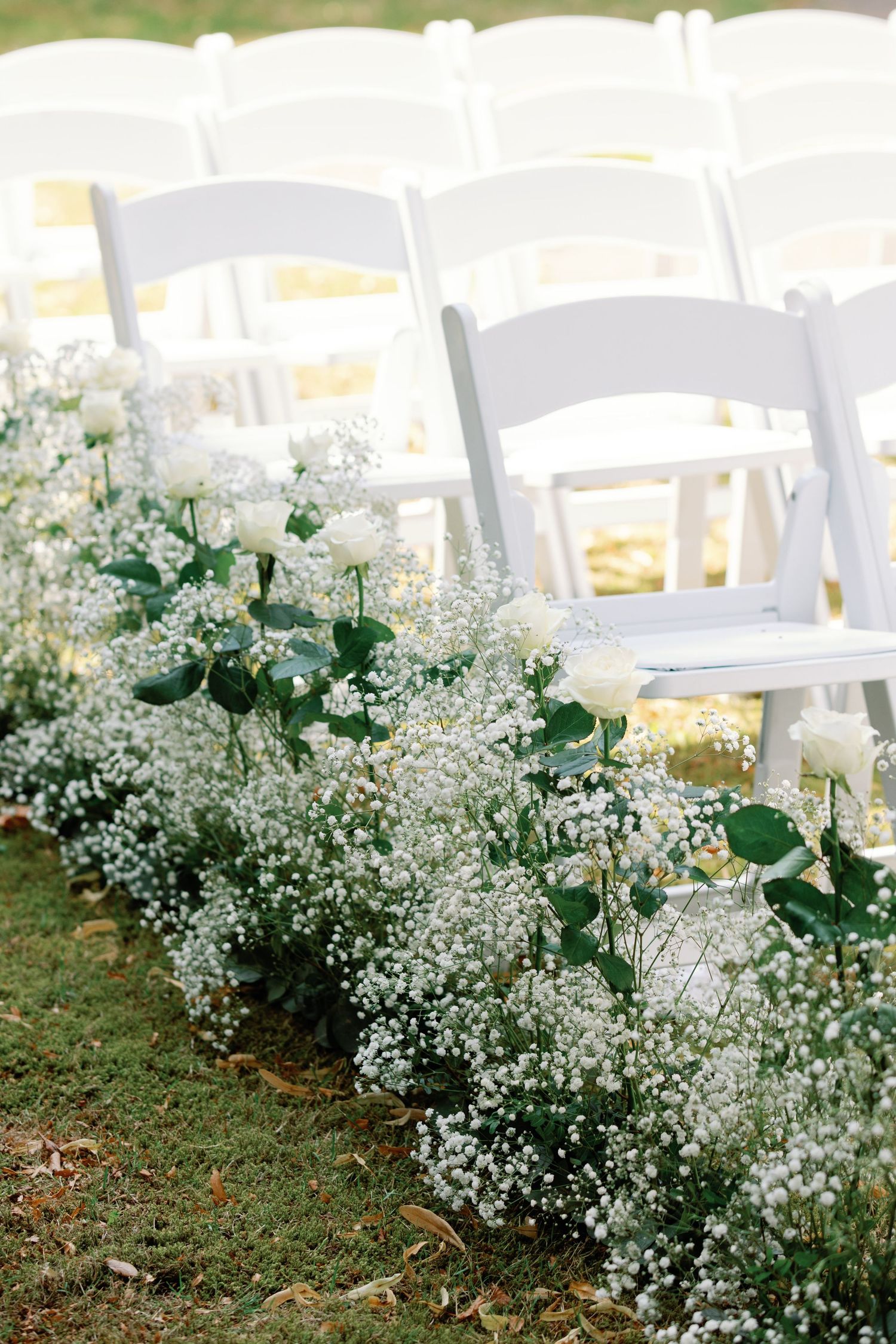 Delicate white baby's breath flowers line a garden path with white chairs in the background for an outdoor wedding ceremony.