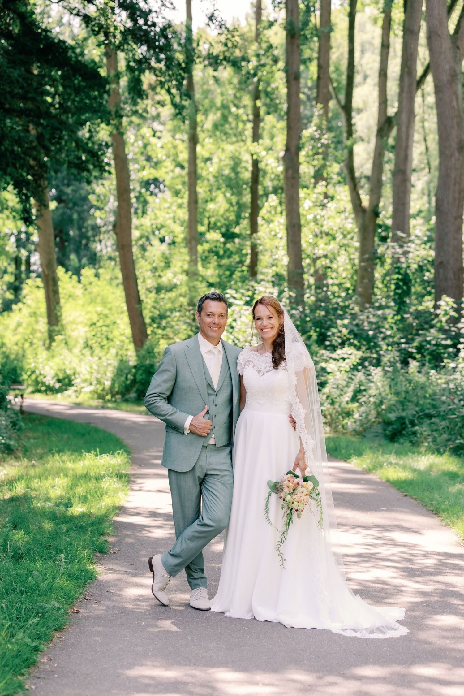 A couple poses together on a sunlit wooded path during their outdoor wedding celebration.
