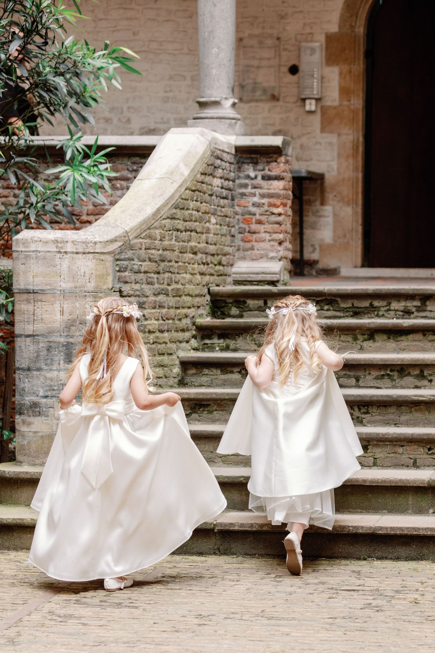 Two figures in flowing white dresses and floral crowns ascending stone steps at a historic building entrance.