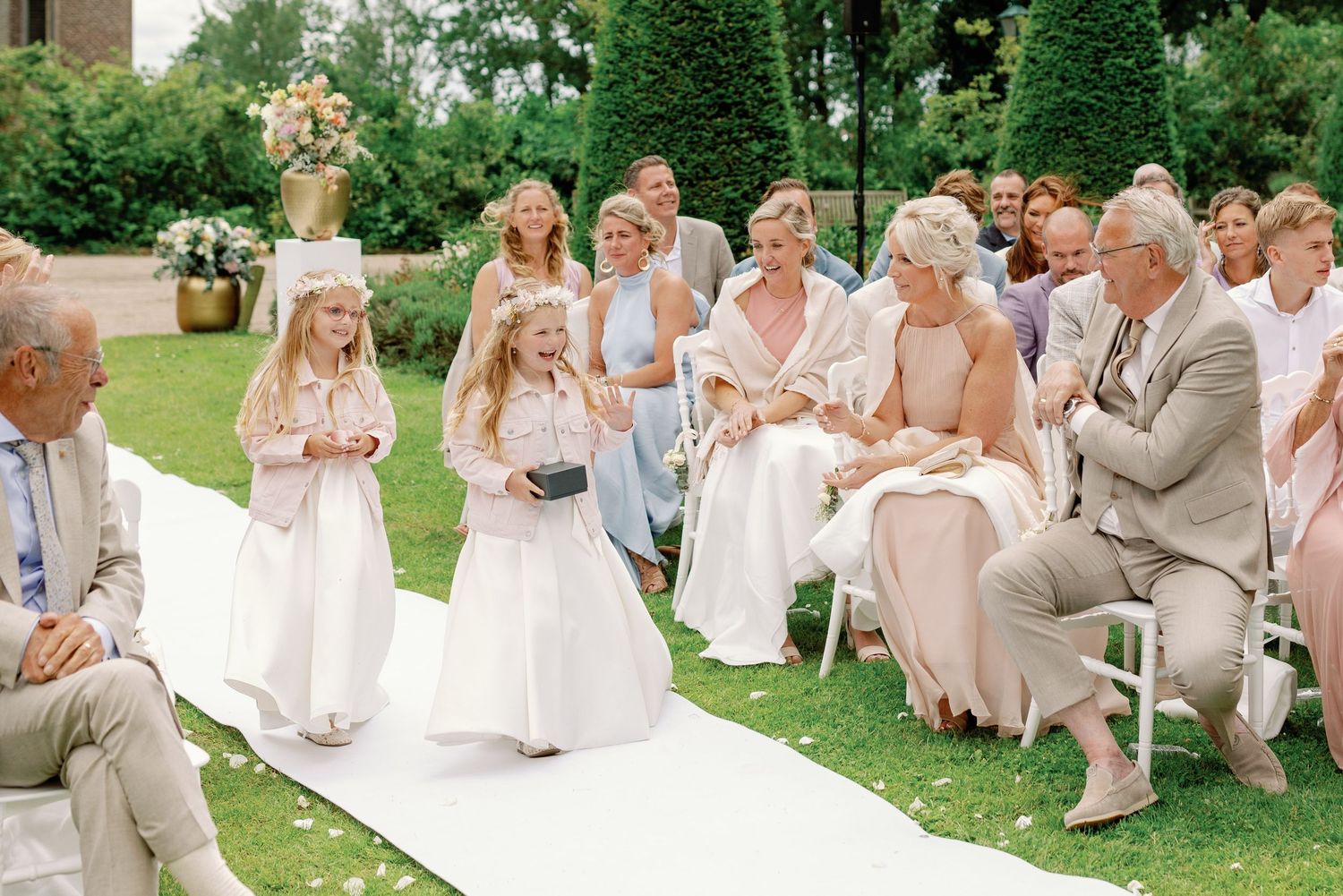 Wedding guests in pastel attire seated on white chairs during an outdoor garden ceremony.