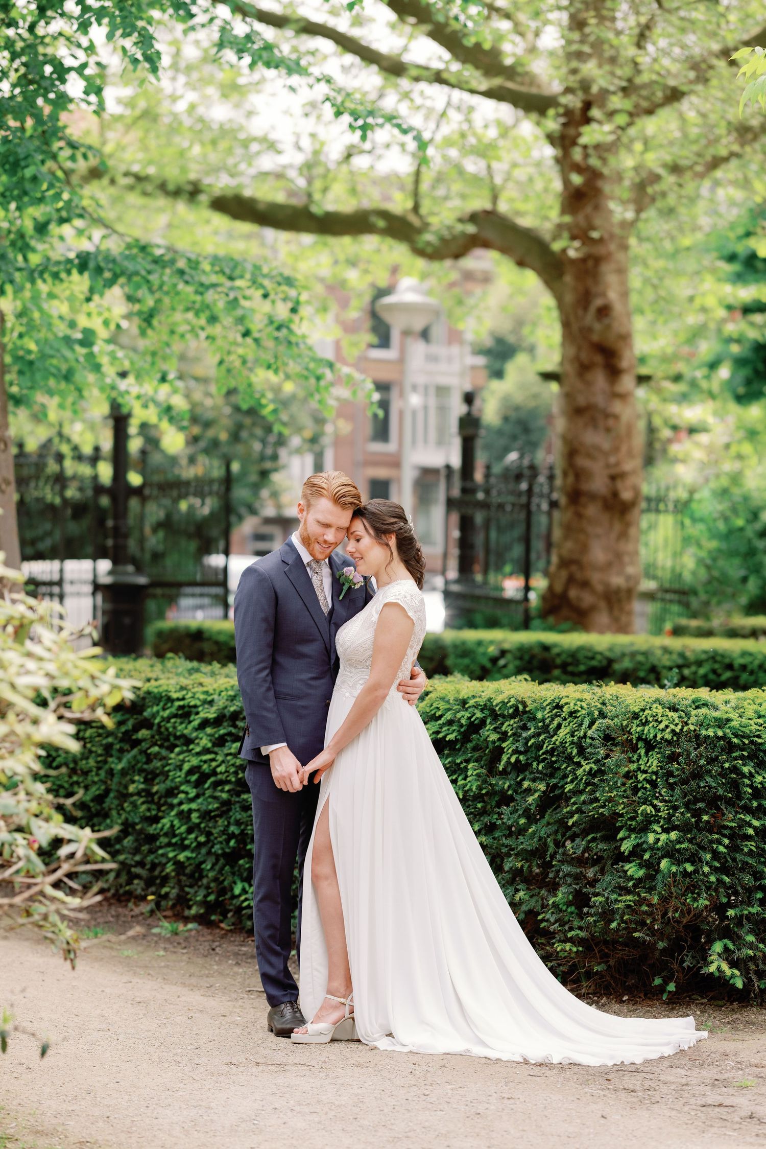 A tender wedding moment captured in a garden setting with trees and manicured hedges in the background.