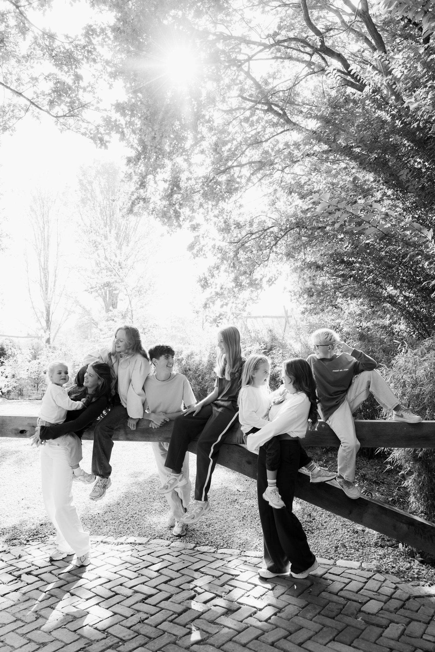 Black and white photo of a family of adults and children sitting and standing along a wooden rail on a brick path under sunlit trees.