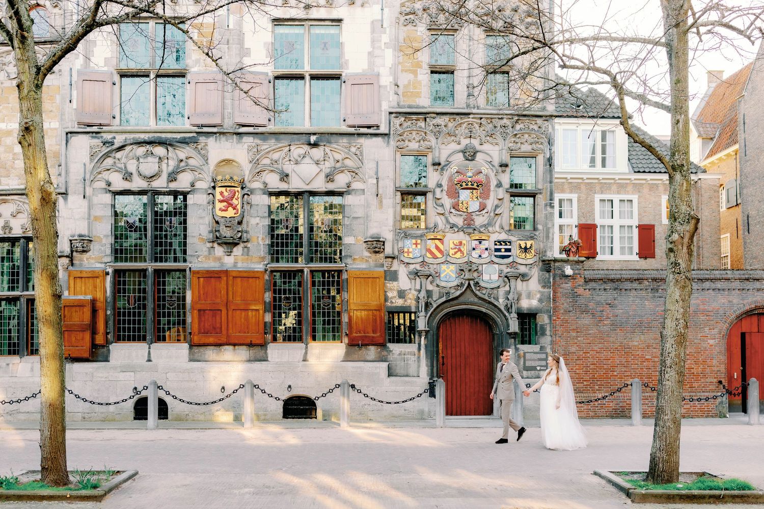 Bride in a white wedding gown and groom walking past an ornate historic Dutch building with red shutters, heraldic crests and bare trees.