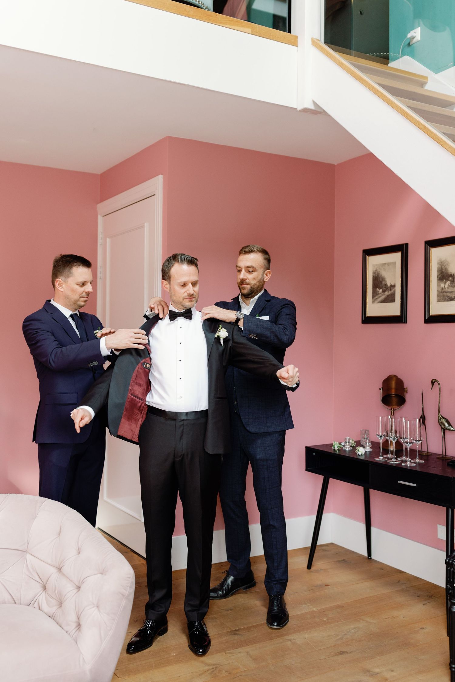 Groomsmen helping groom into tuxedo jacket in pink room before wedding ceremony.