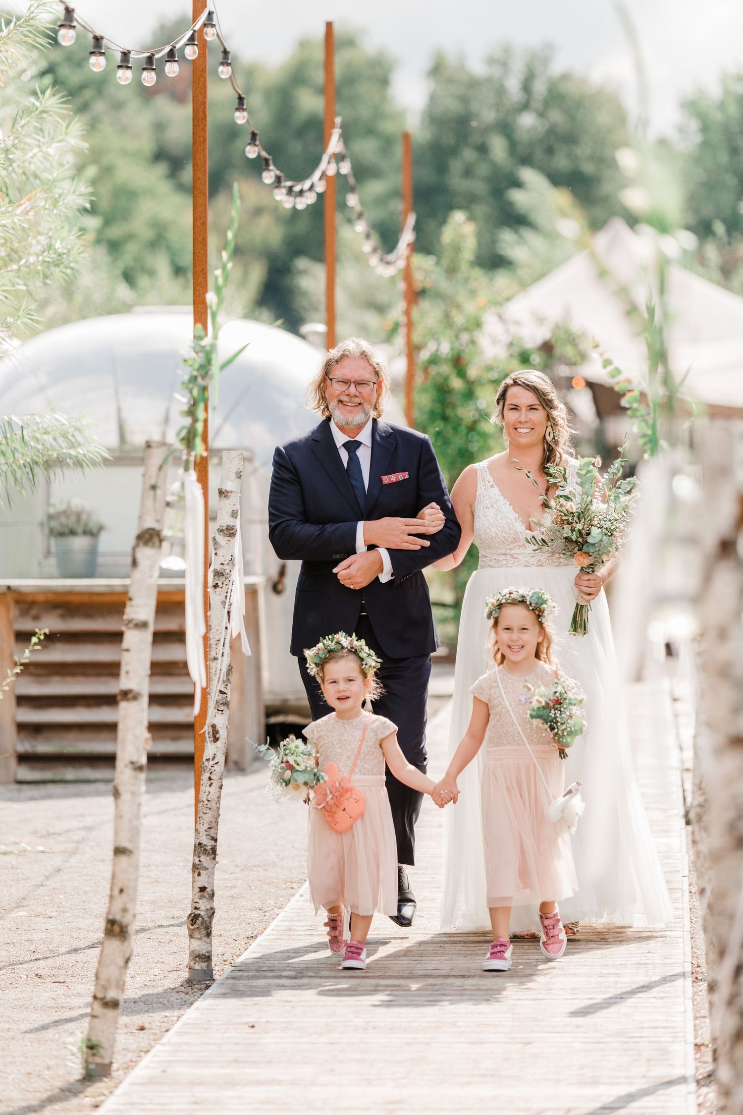 A wedding party walks down a decorated pathway with birch poles and string lights in a garden setting.