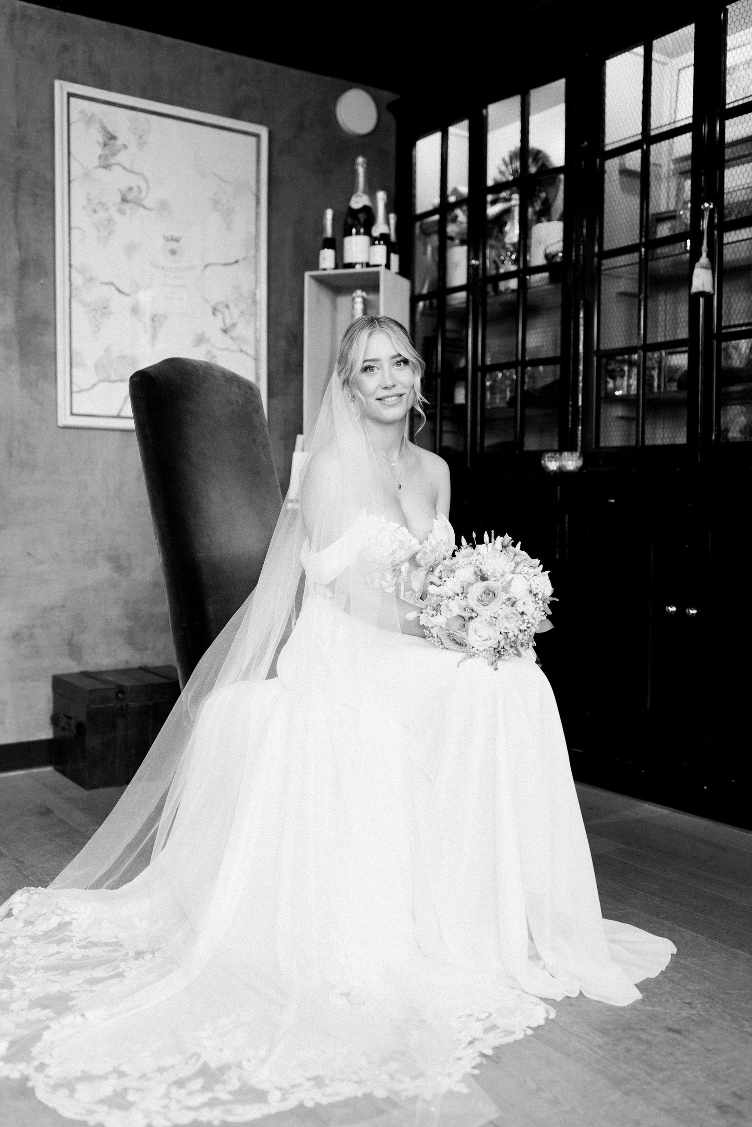 A bride in an elegant white gown poses in a room with large windows and vintage decor.