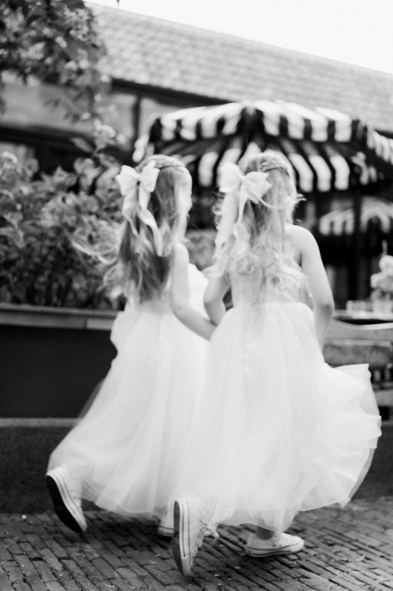 Young flower girls in flowing white dresses twirl and dance together on cobblestone pavement at an outdoor event.