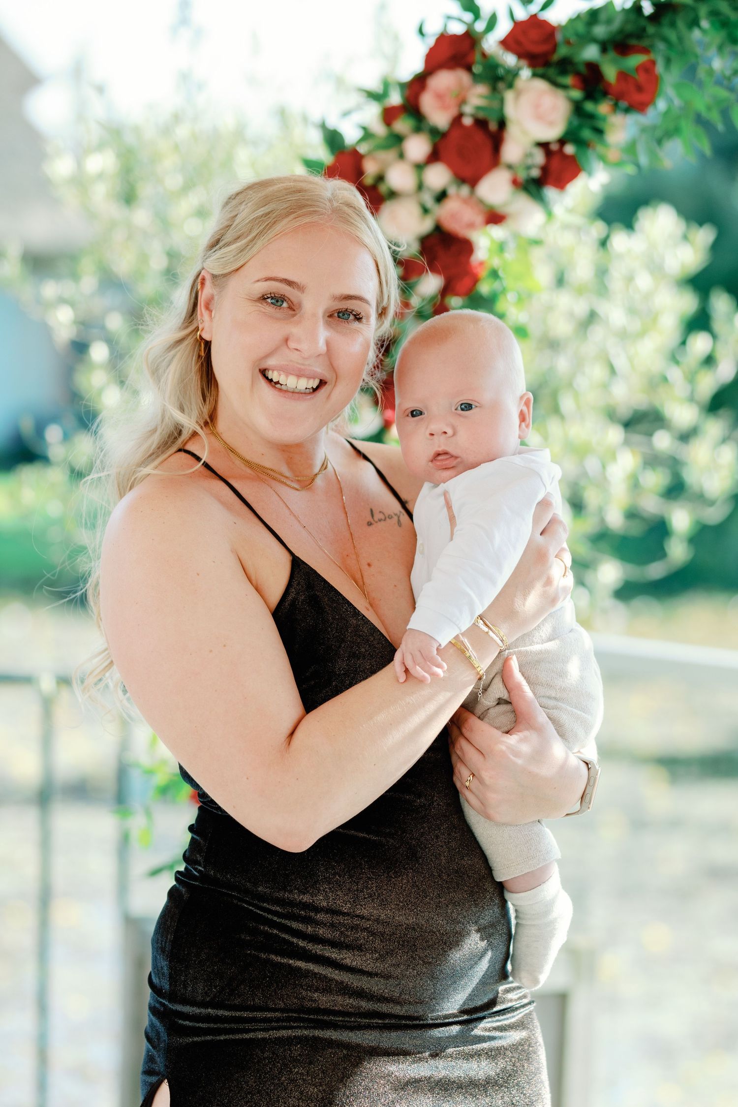 A woman in a black dress holds and smiles at a baby dressed in white.
