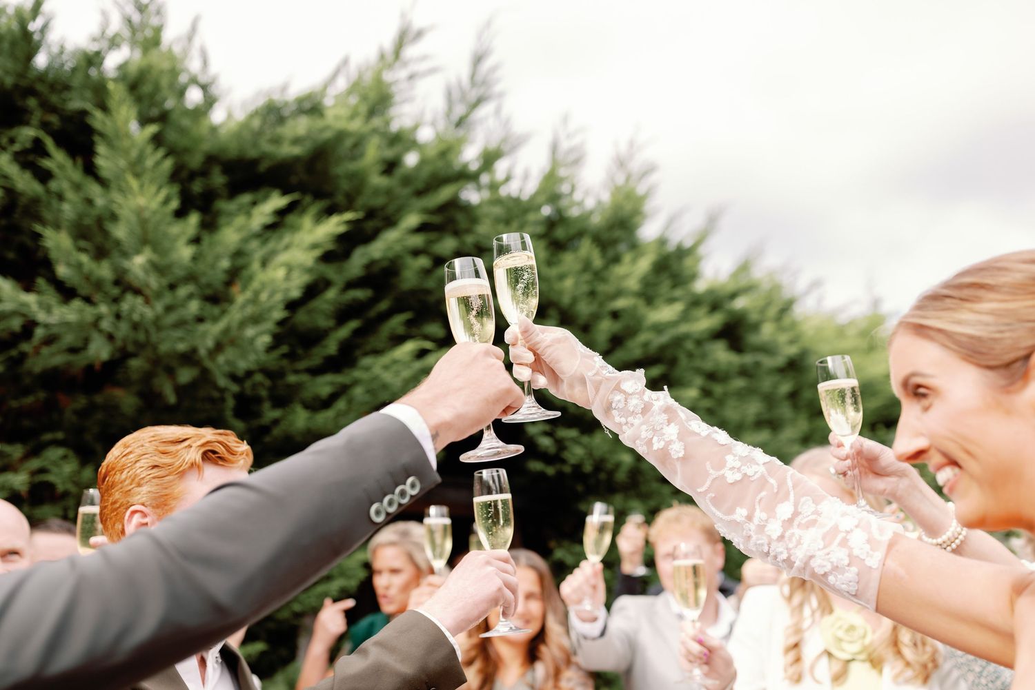 A group raises champagne glasses in a celebratory toast outdoors with evergreen trees in the background.