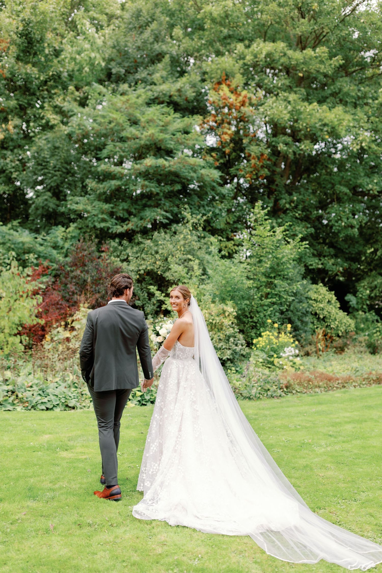 A couple walks through a lush green garden setting with a flowing white wedding dress trailing behind them.