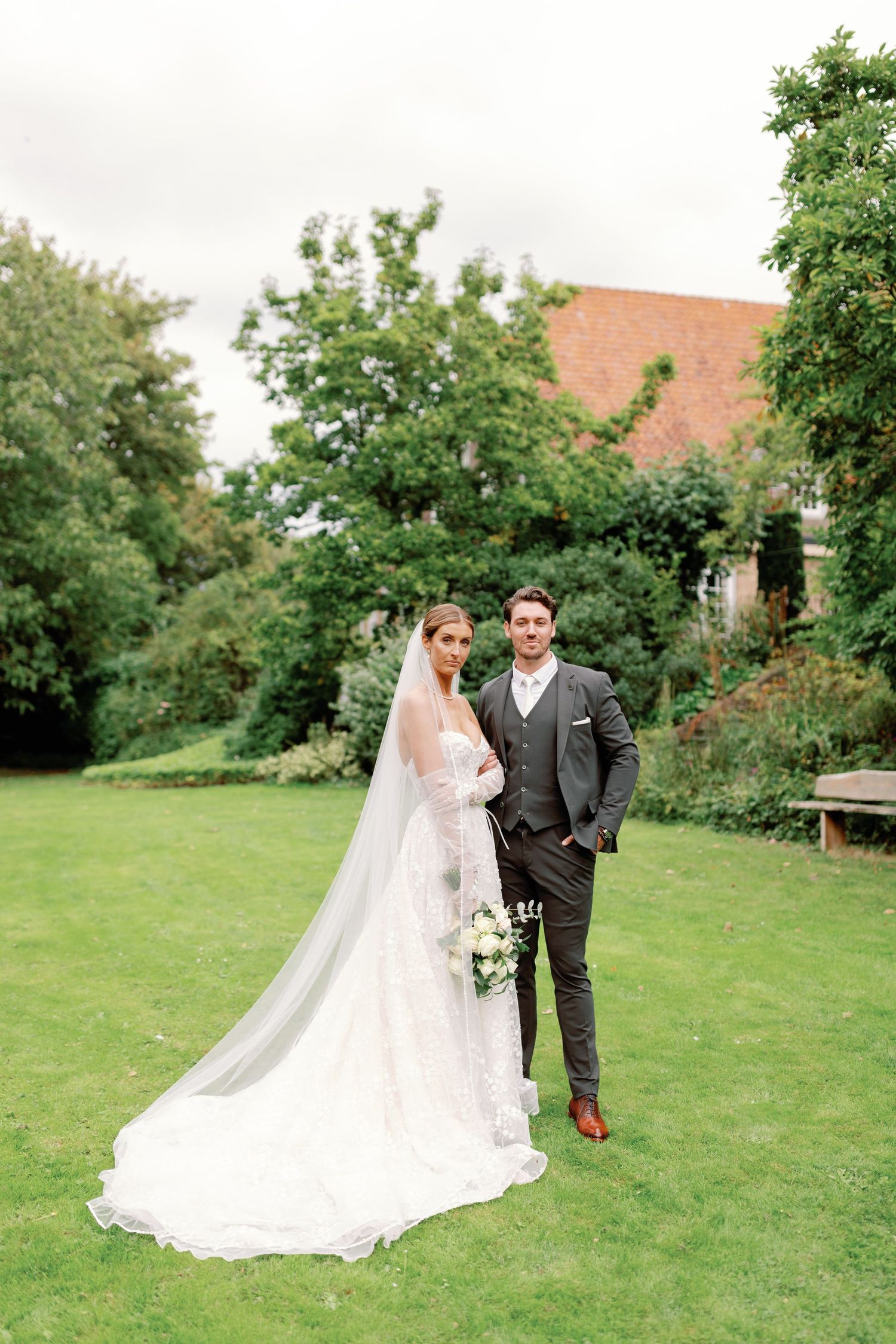 A couple stands on a lush green lawn surrounded by trees and gardens on their wedding day.