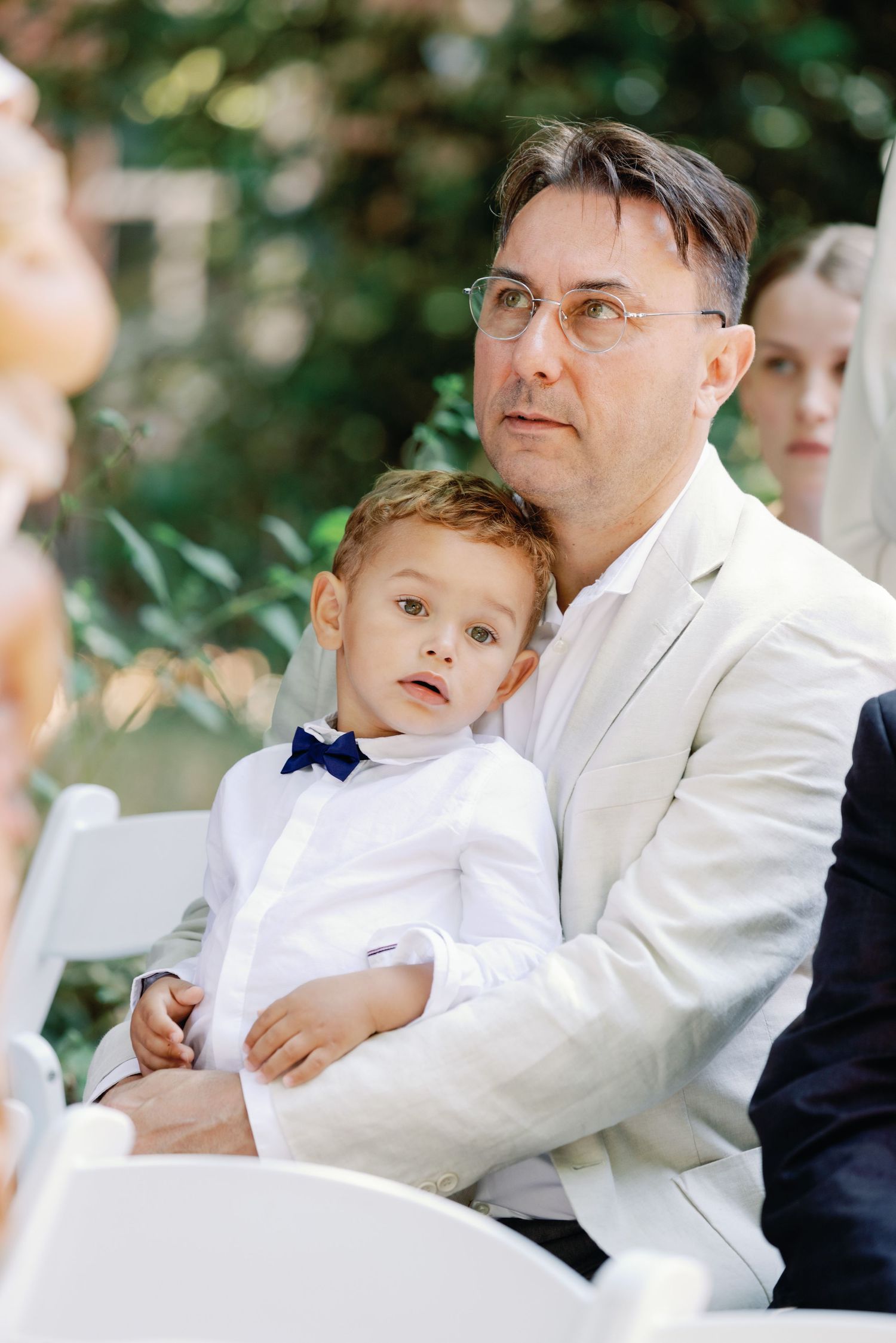 Young ring bearer in white suit sitting on outdoor chairs during a wedding ceremony.