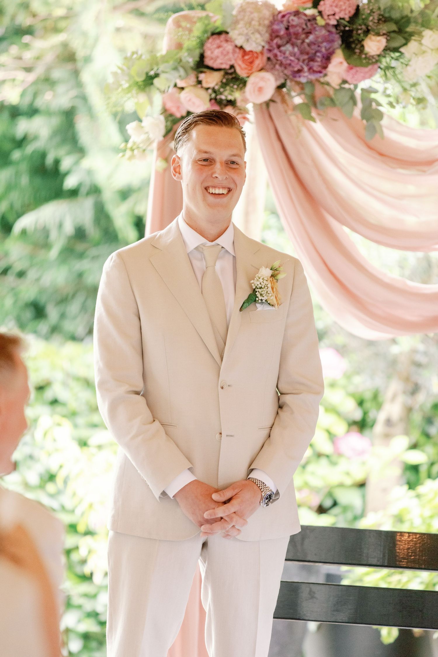 Wedding party members in light beige suits stand in a row during an outdoor ceremony with pink floral decorations.