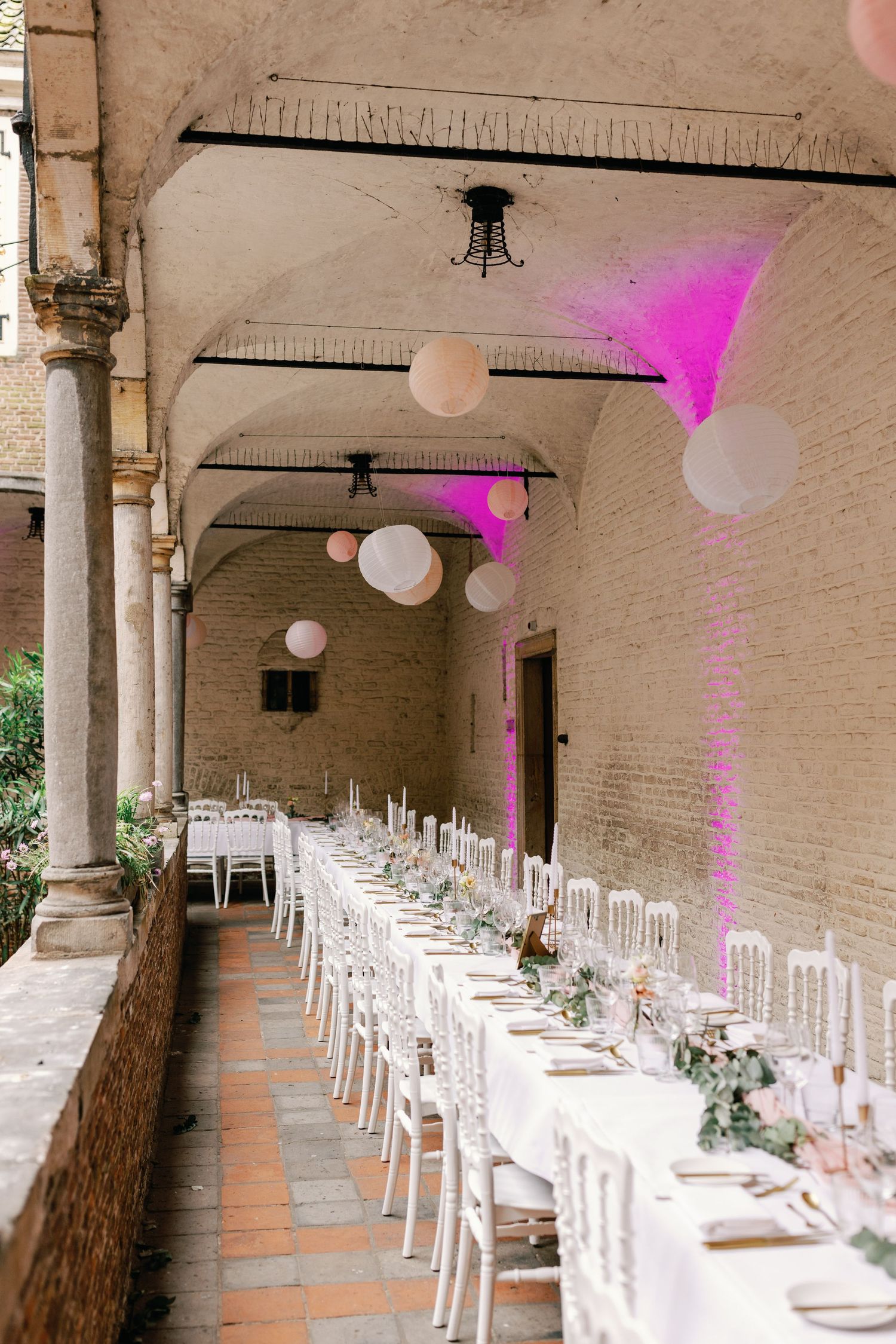 Long wedding reception table setup under arched columns with white lanterns and pink accent lighting in historic venue.