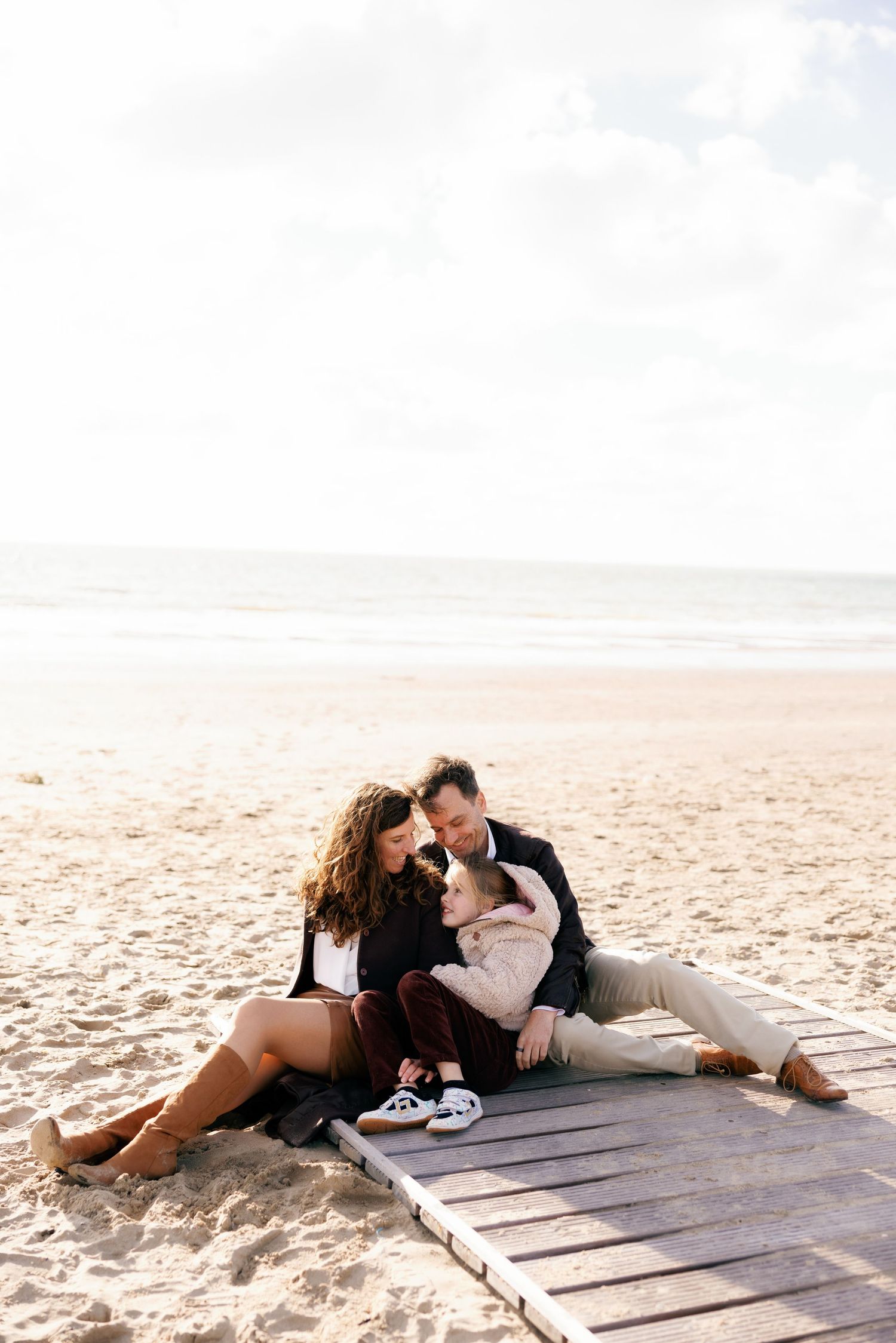 Family of three sitting on a wooden boardwalk on a sandy beach, sharing a tender moment together.