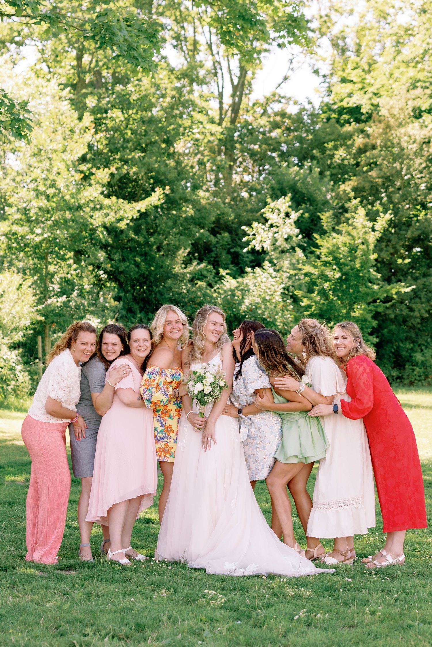 A wedding party in pink and white dresses poses playfully in a series of outdoor photos on a grassy lawn with trees.