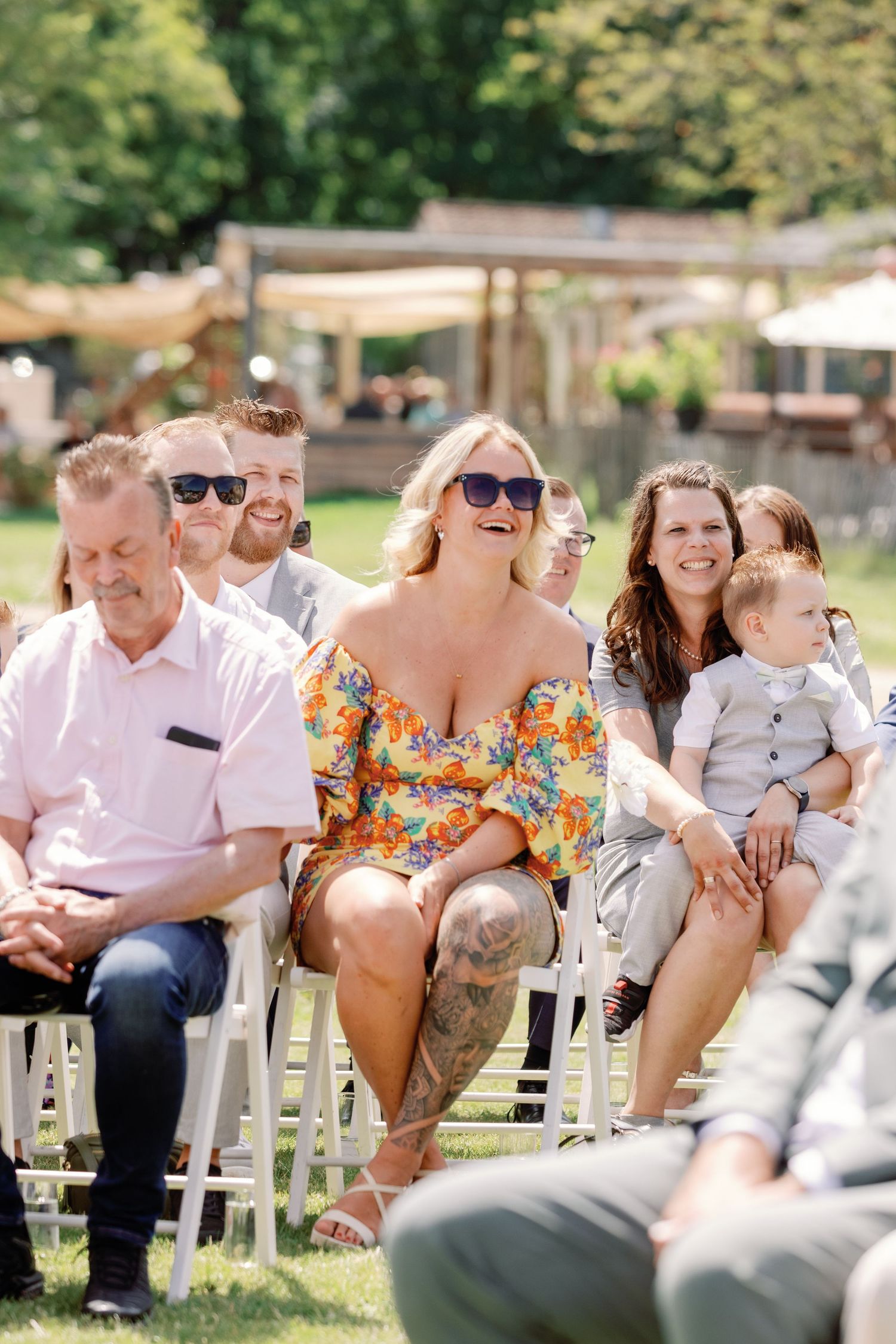 Wedding guests share laughter during an outdoor ceremony while seated in white chairs on a sunny day.