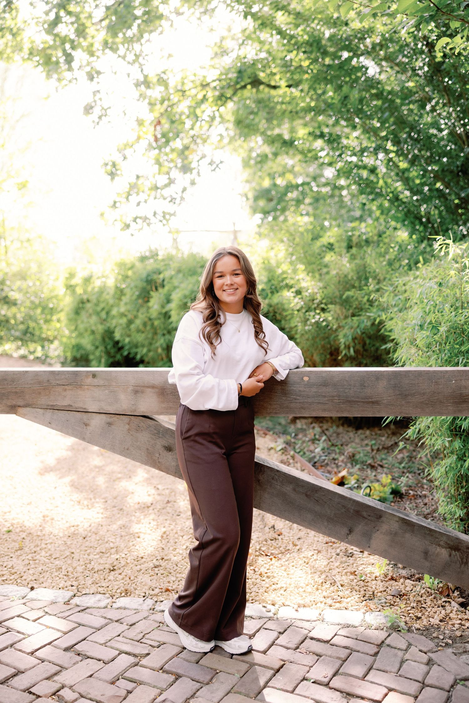 A young woman in a white blouse and brown trousers leans on a wooden fence beside a brick path with lush greenery behind her.