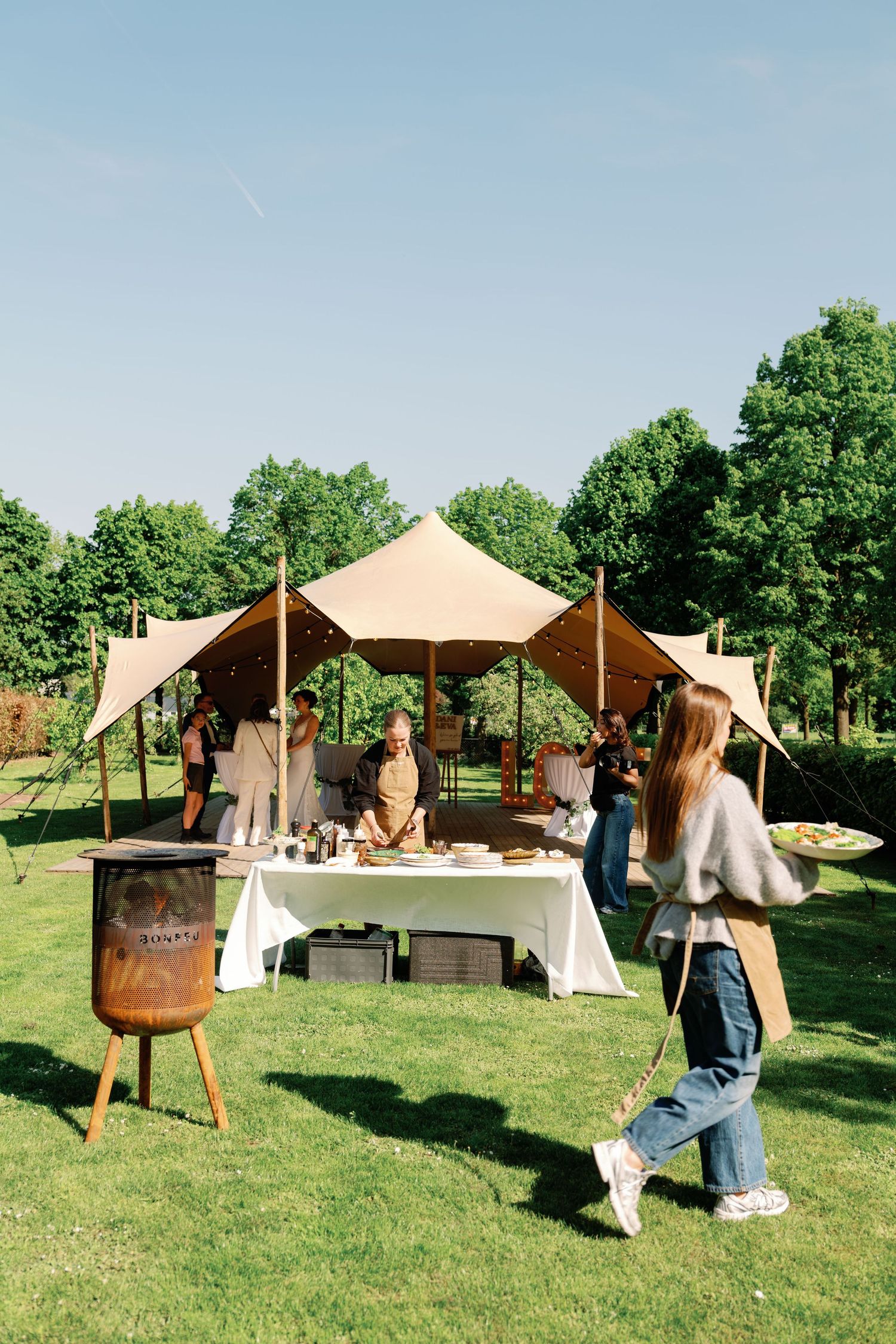 Outdoor event setup with beige stretch tent and vendor table on green lawn during summer day with trees in background.